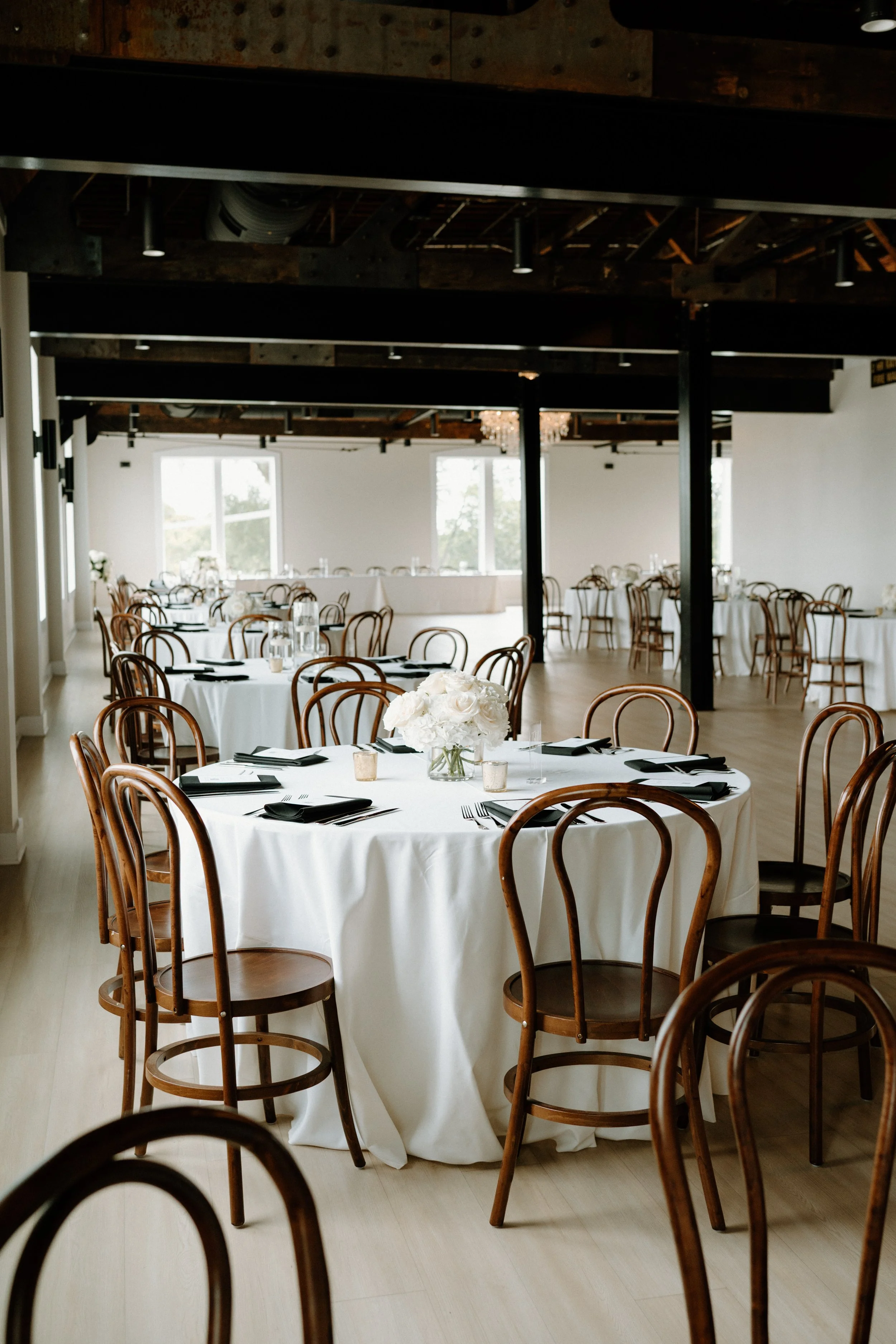Elegant banquet hall with round tables covered in white tablecloths, decorated with white floral centerpieces, black napkins, and candles, set for a formal event.
