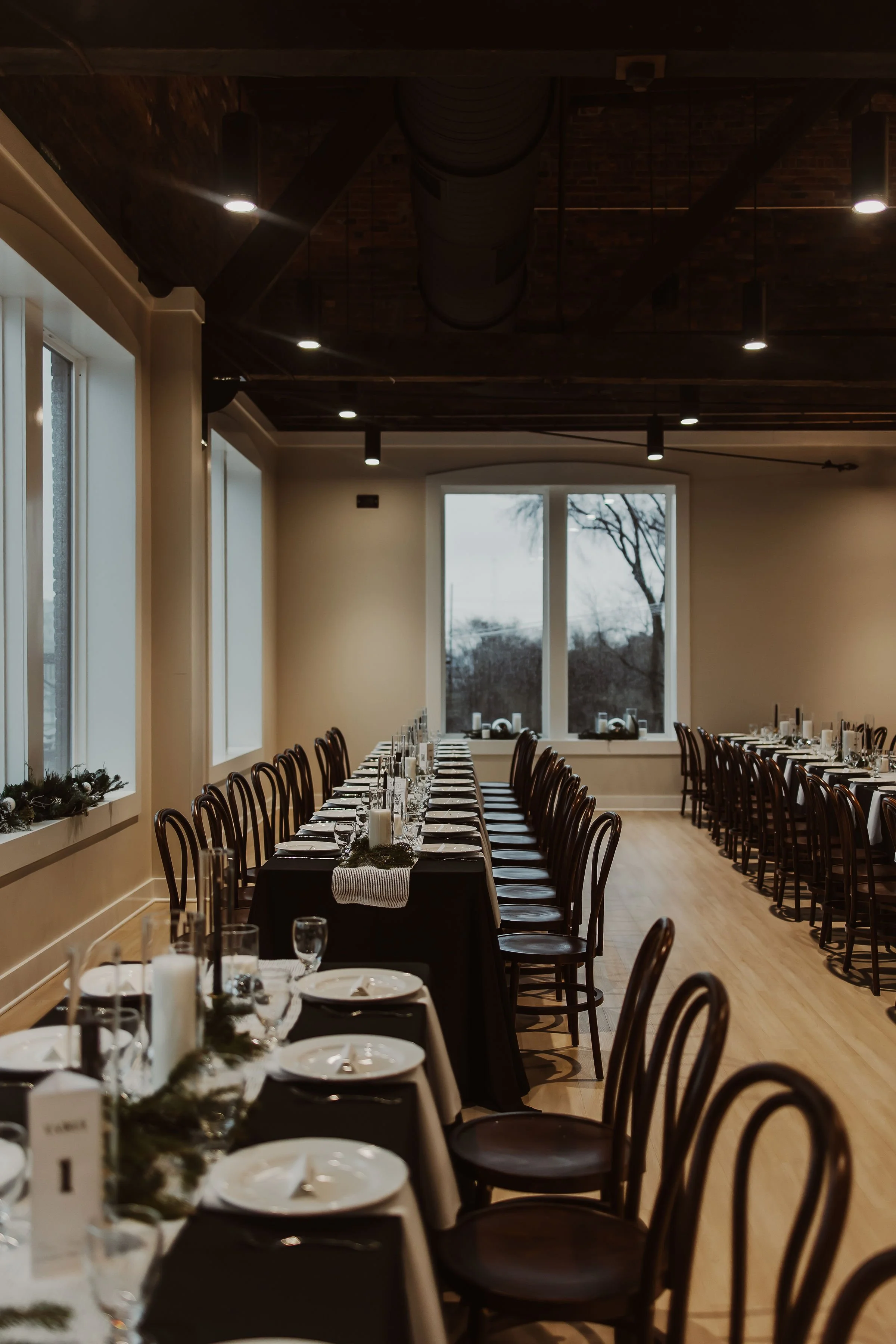 Elegant banquet hall with long tables set for a meal, decorated with candles and greenery, large windows, dark wood chairs, and warm lighting.