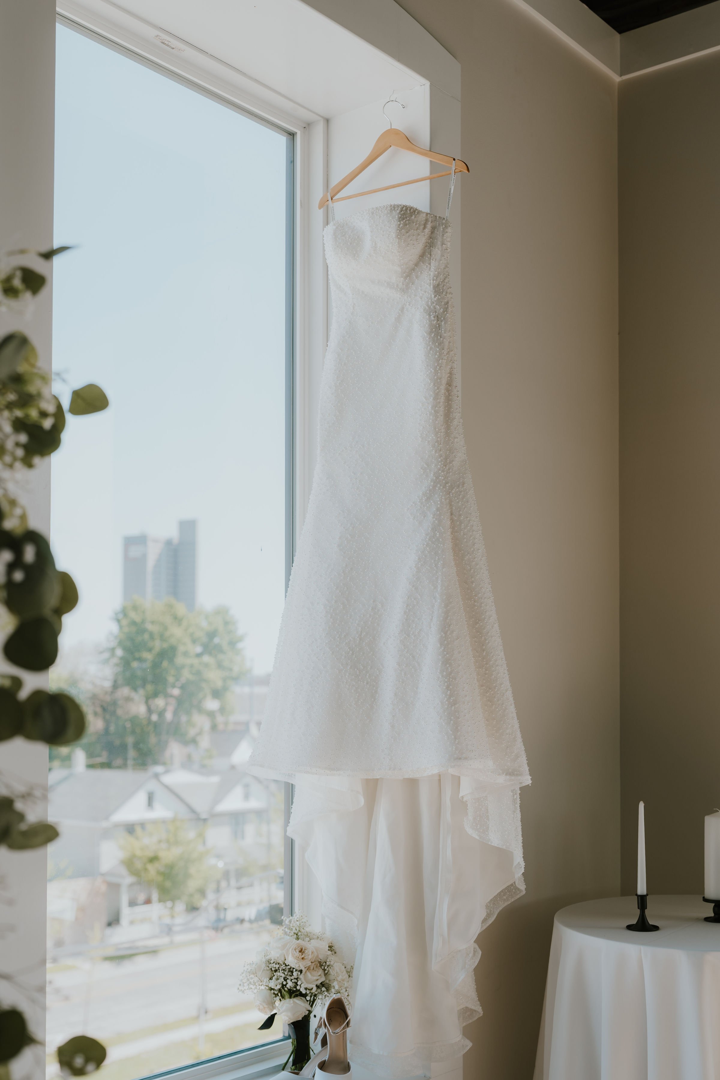 A wedding dress hanging on a wooden hanger near a large window, with a bouquet of white flowers and a pair of beige shoes at the base.