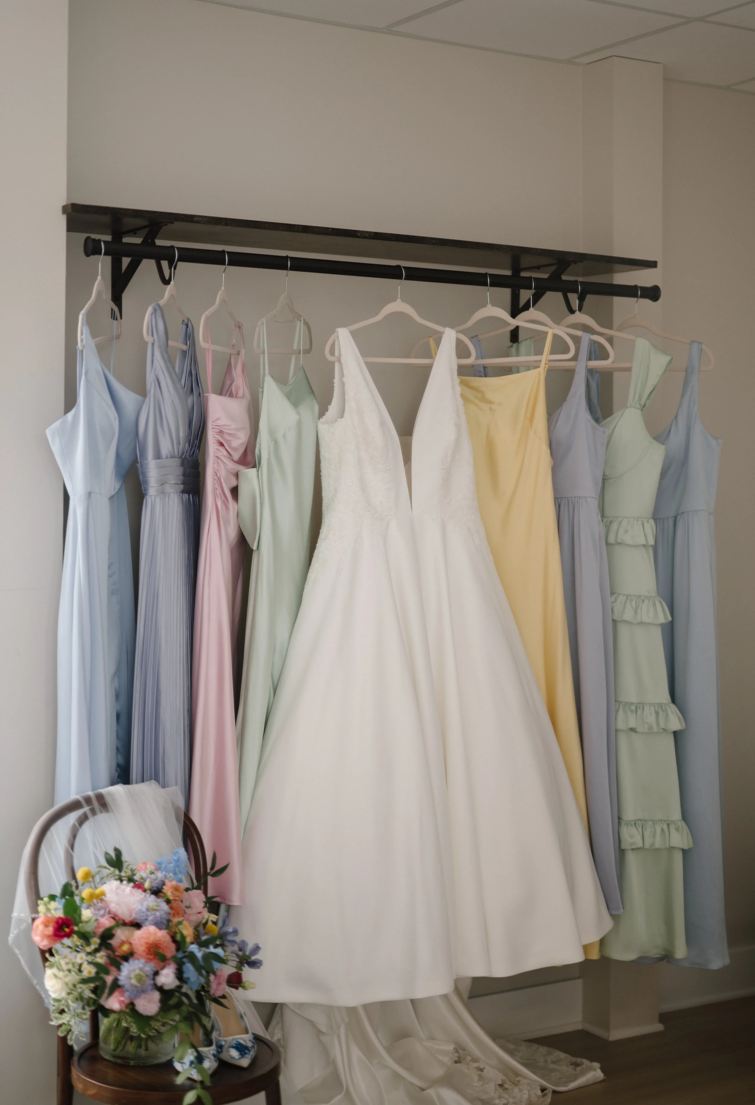 Several pastel-colored dresses hanging on a black clothing rack with a white wedding gown in front, complemented by a bouquet of colorful flowers on a small wooden table.