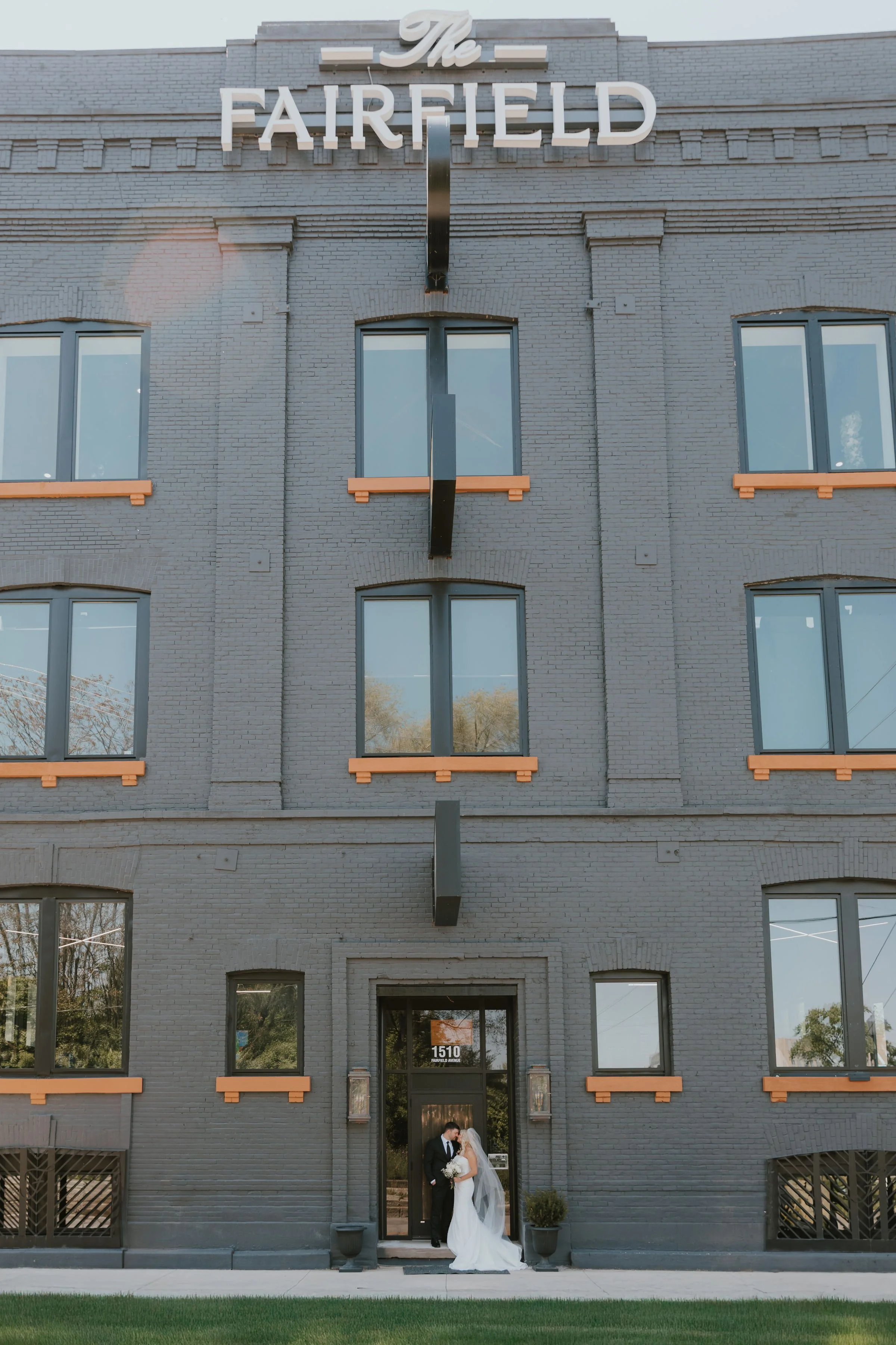A bride and groom standing outside the entrance of The Fairfield, a multi-story brick building, on their wedding day.