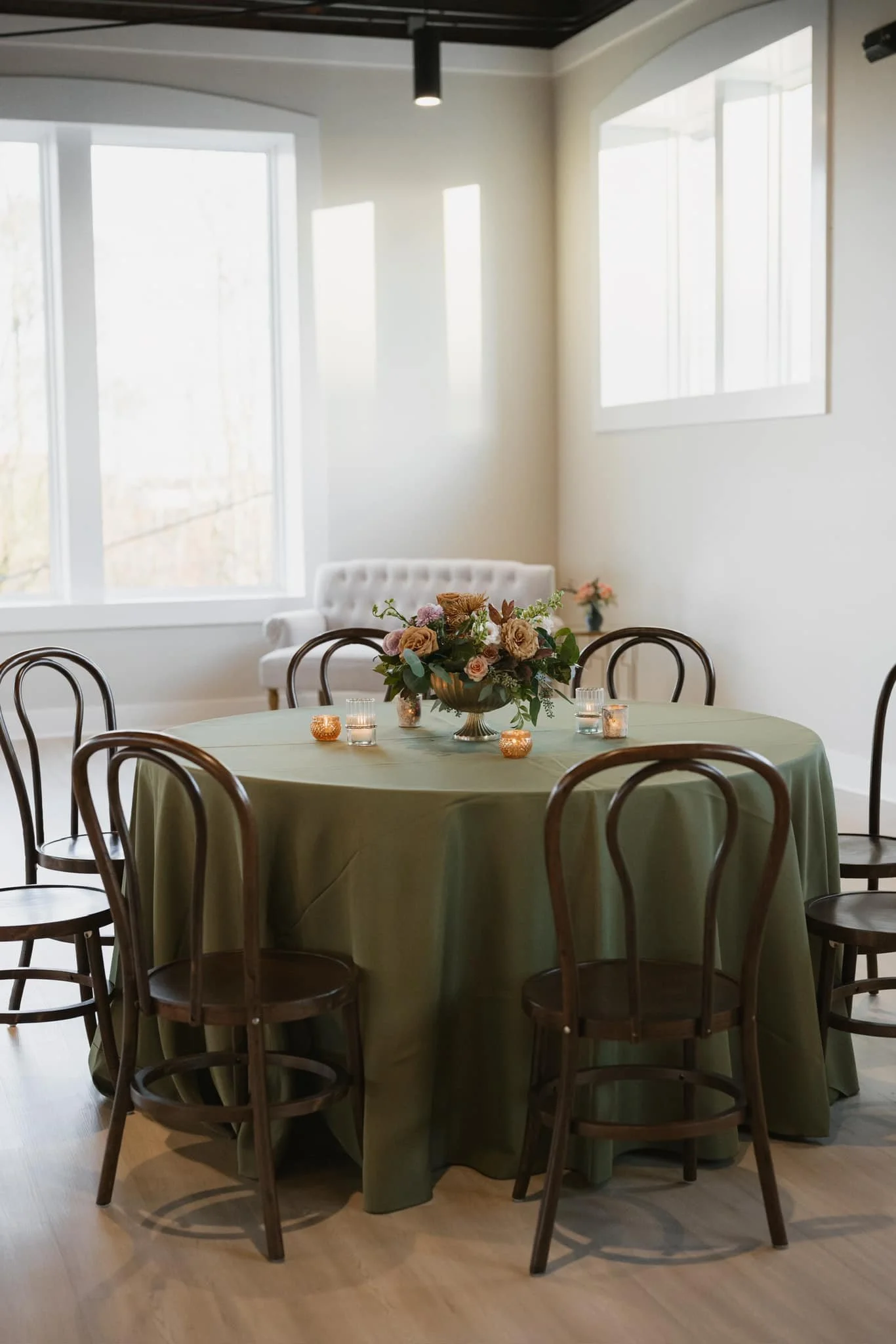 A round table draped with a green tablecloth, decorated with a floral centerpiece and small candles, surrounded by wooden chairs in a well-lit room with large windows.