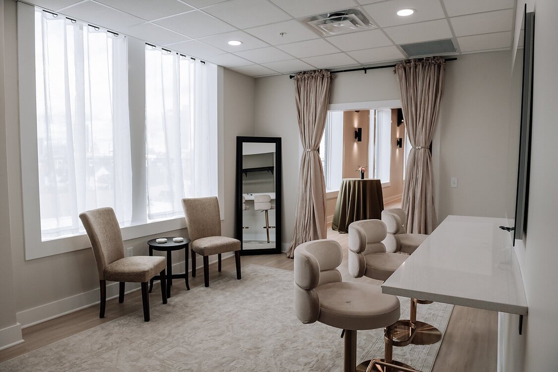 A bright room with beige curtains, two beige chairs near the window, a black framed mirror, a white bar-height table with cream-colored barstools, and beige carpeting.