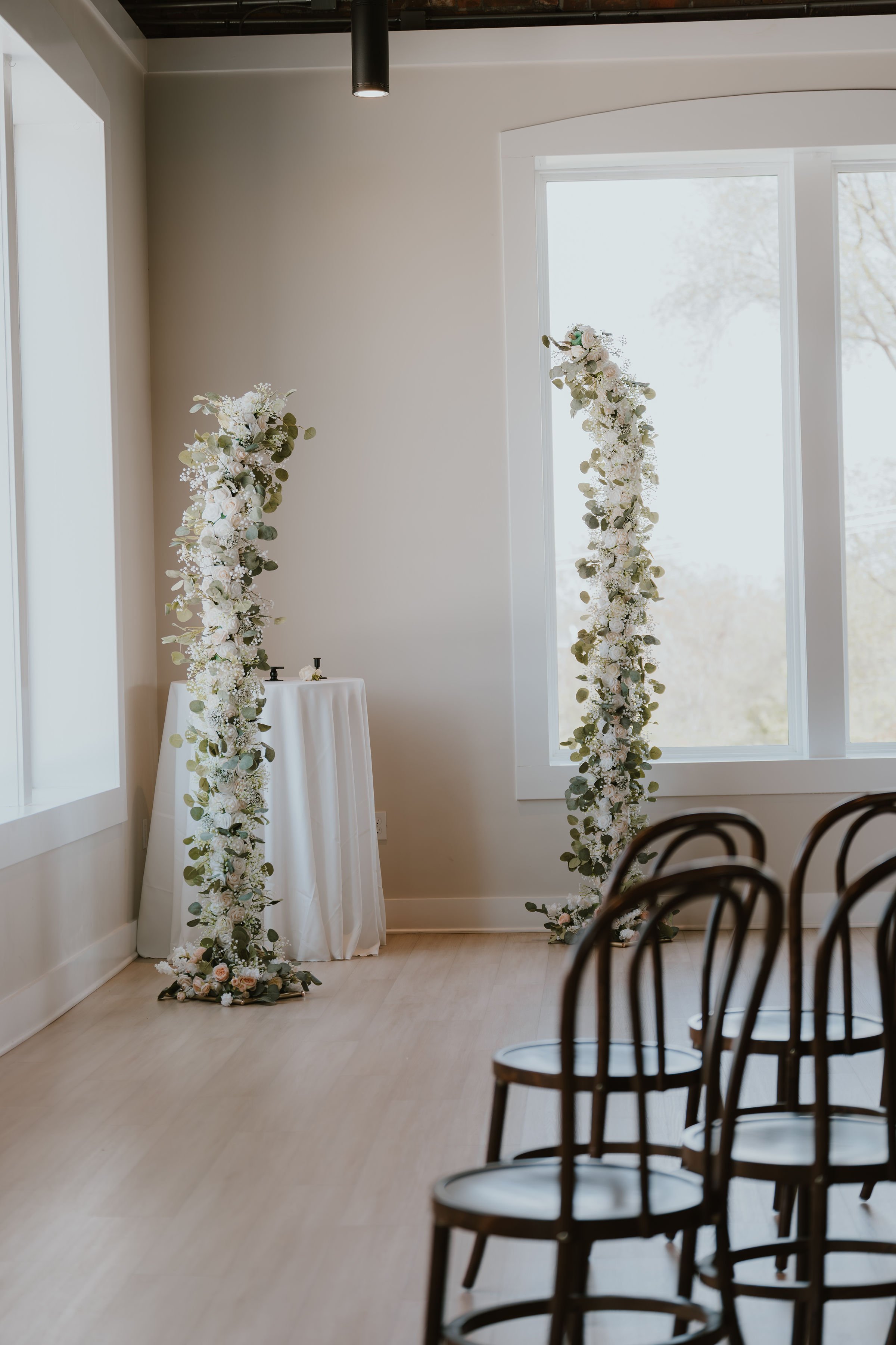 Wedding ceremony setup with two tall floral arches decorated with white flowers and green leaves, positioned near a large window.