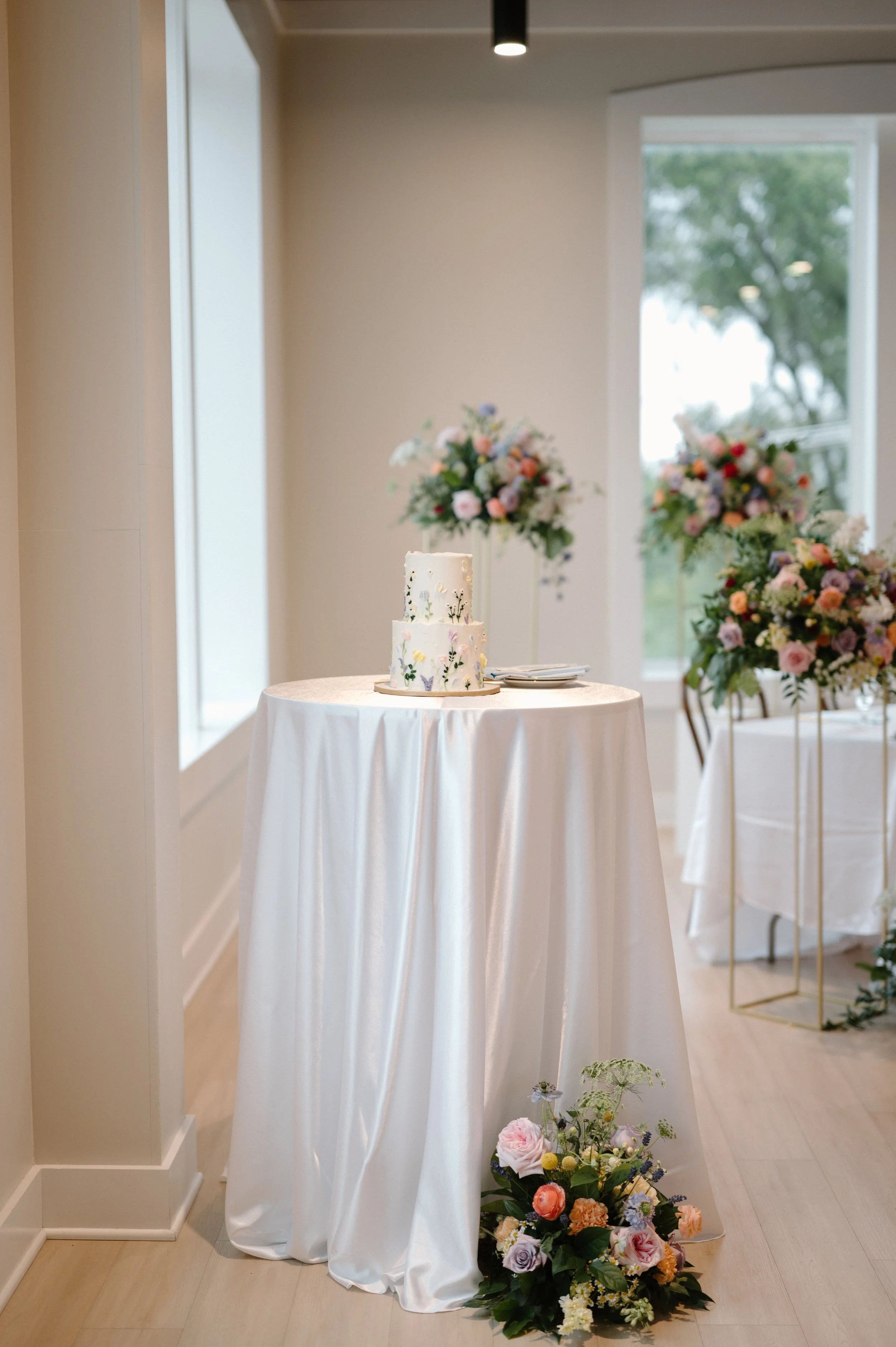 A wedding cake on a tall, round table with a white satin tablecloth, decorated with a floral cake topper. Behind the table, there are large floral arrangements on stands, with pink, purple, white, and orange flowers in a bright, elegant room with lar