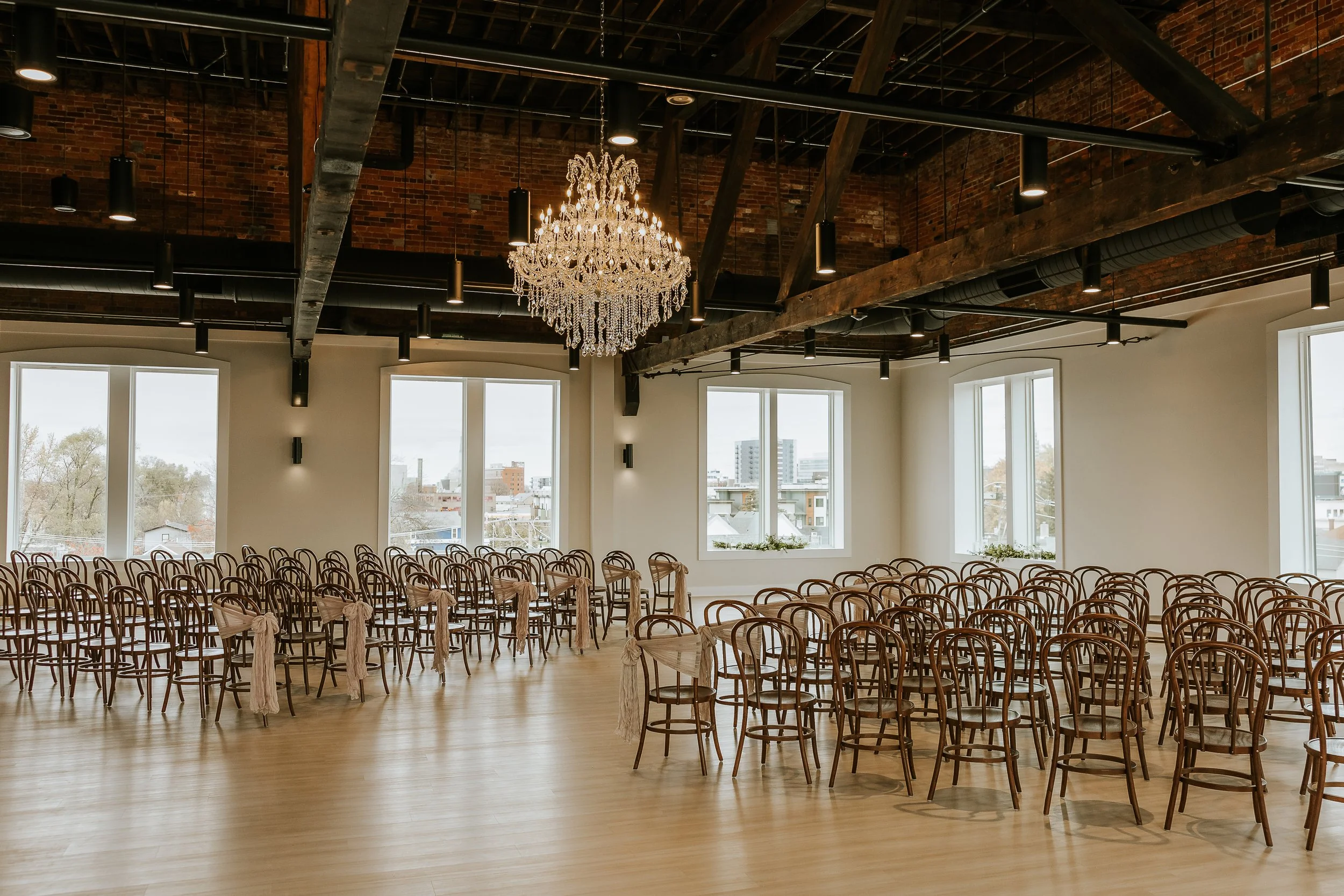 Empty event space with wooden chairs adorned with beige sashes, large windows, exposed brick and ceiling beams, and a chandelier hanging from the ceiling.