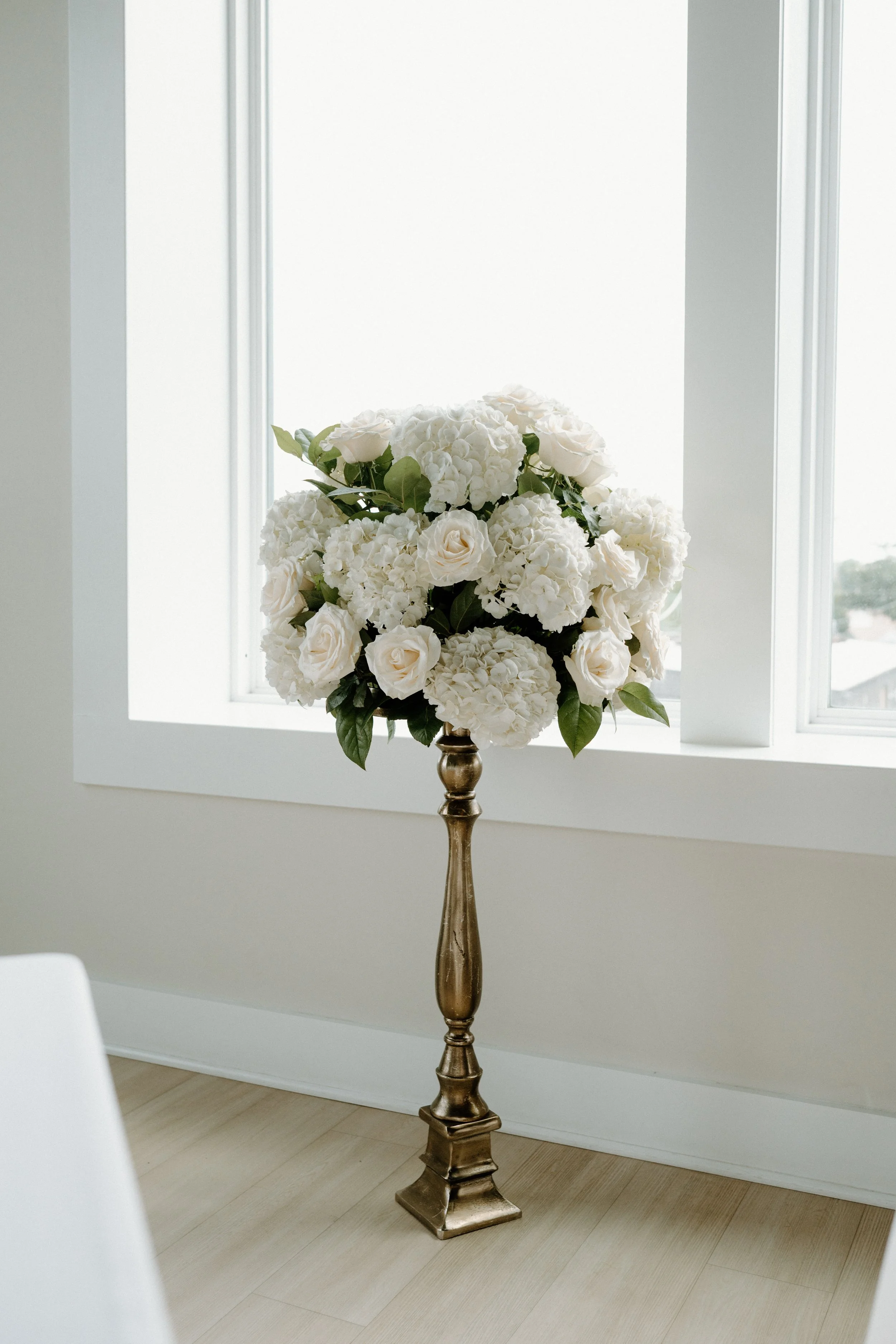 Elegant floral arrangement of white roses and hydrangeas on a tall ornate gold stand, set against a bright window in a minimalist room.