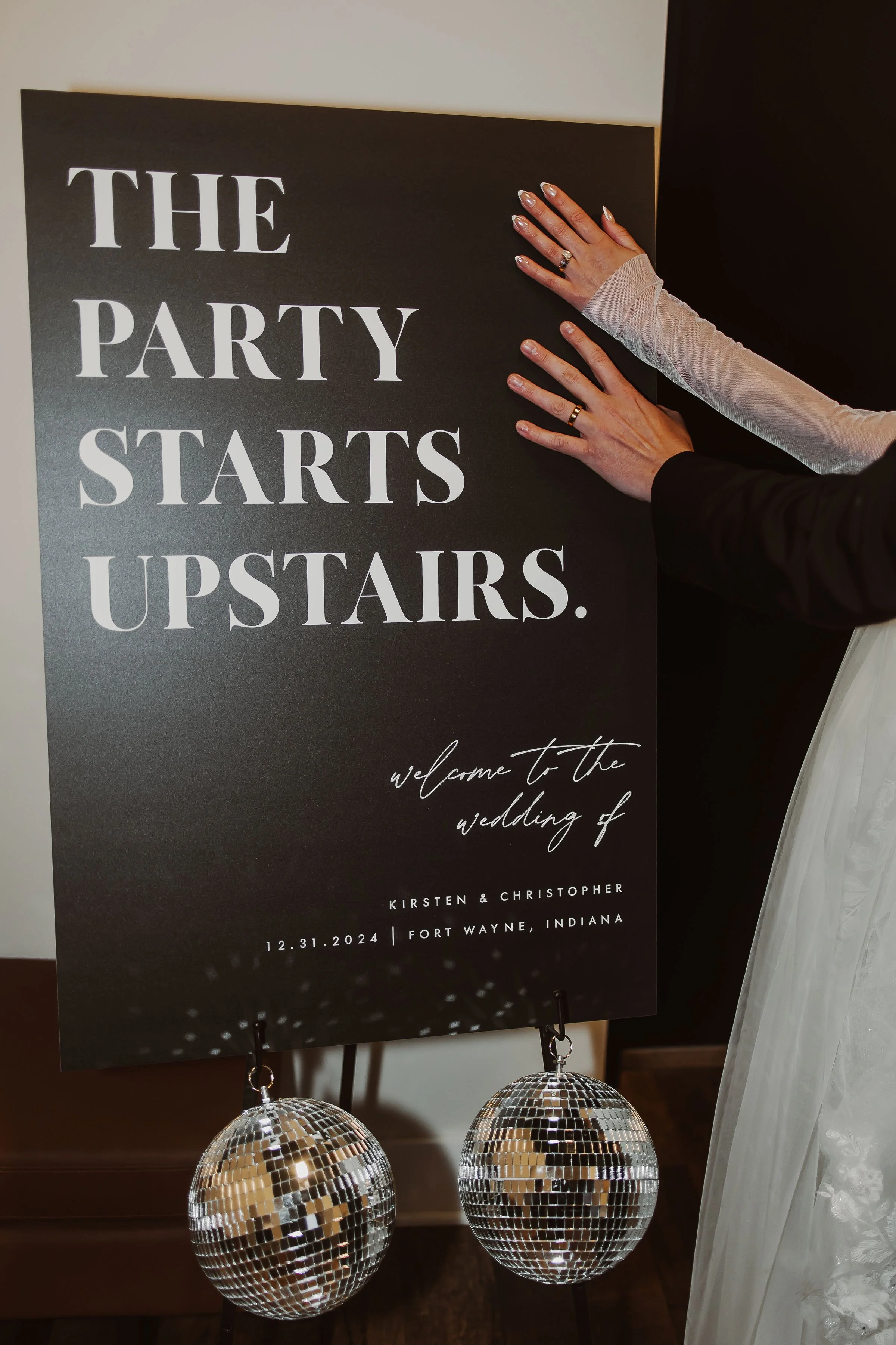 A sign at a wedding reception that says 'The party starts upstairs. Welcome to the wedding of Kristen & Christopher, 12.31.2024, Fort Wayne, Indiana.' There are two disco balls hanging at the bottom of the sign, and a person is adjusting the sign wit
