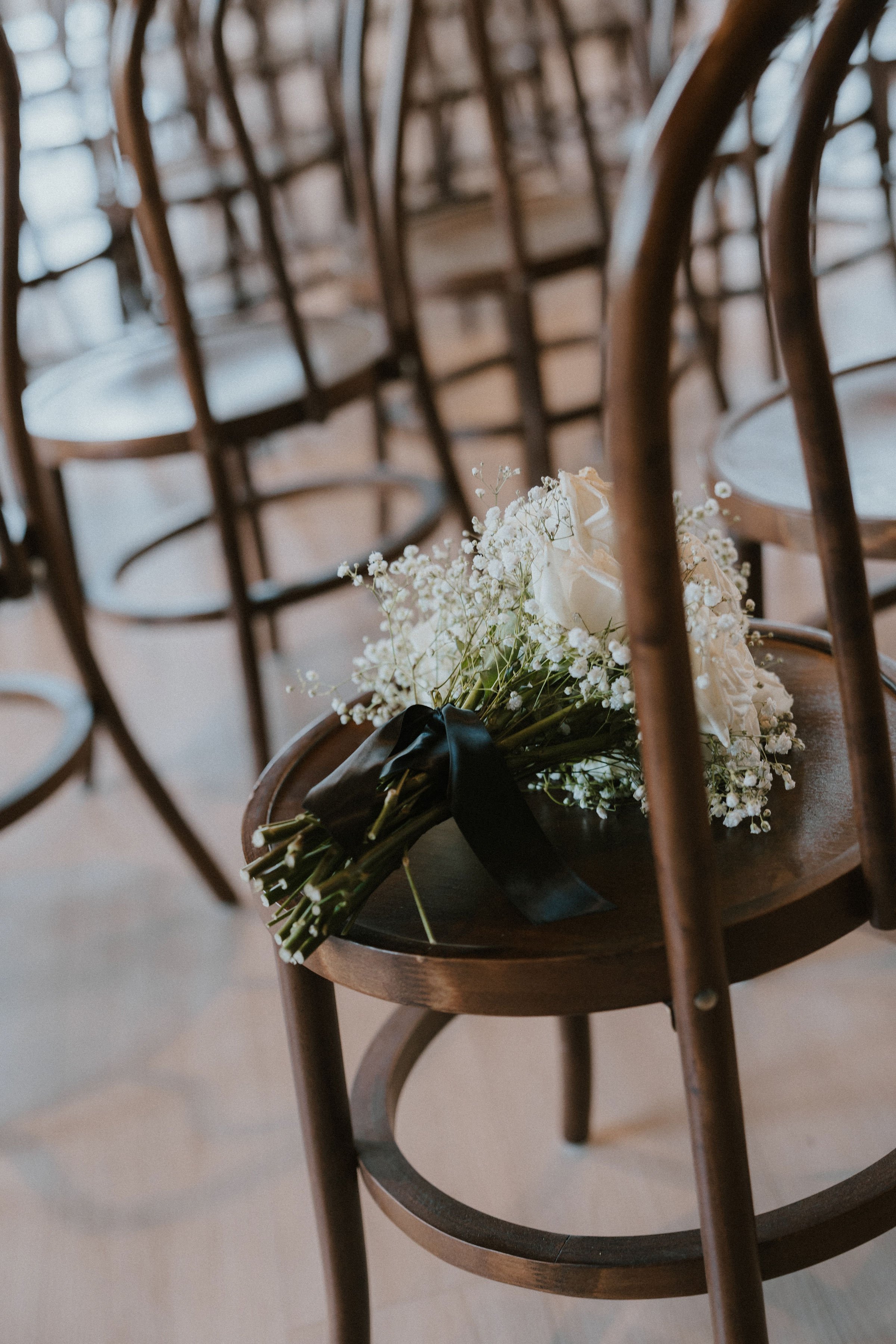 A bouquet of white roses and baby's breath with a black ribbon, resting on a wooden chair surrounded by others in a row.