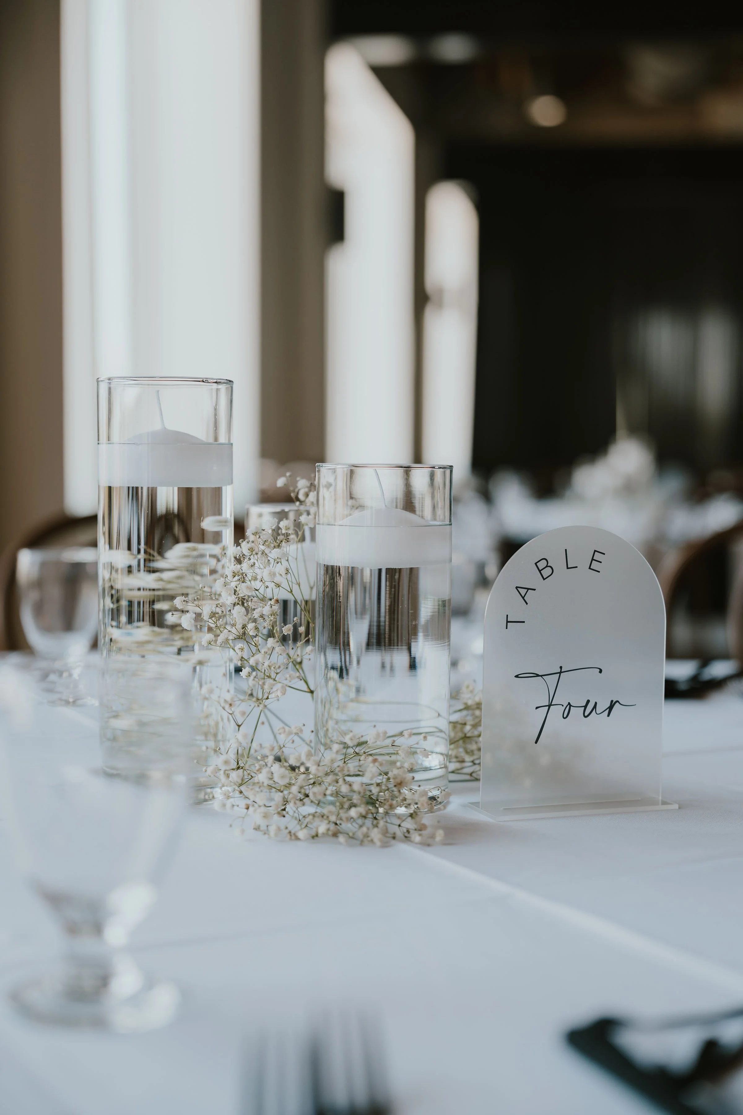 Elegant table centerpiece with two tall cylindrical glass vases filled with water and floating candles, surrounded by delicate white baby's breath flowers, next to a table number sign reading 'Table Four' in cursive font, set on a white tablecloth at