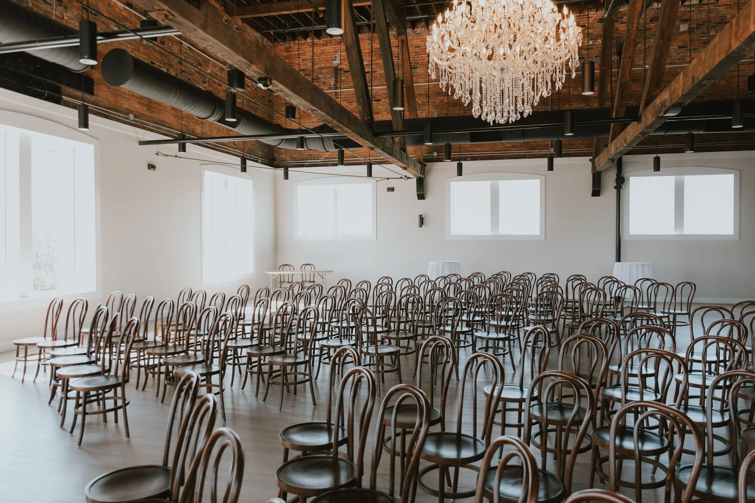 Empty event space with rows of wooden chairs, large windows, exposed brick ceiling, and a chandelier.