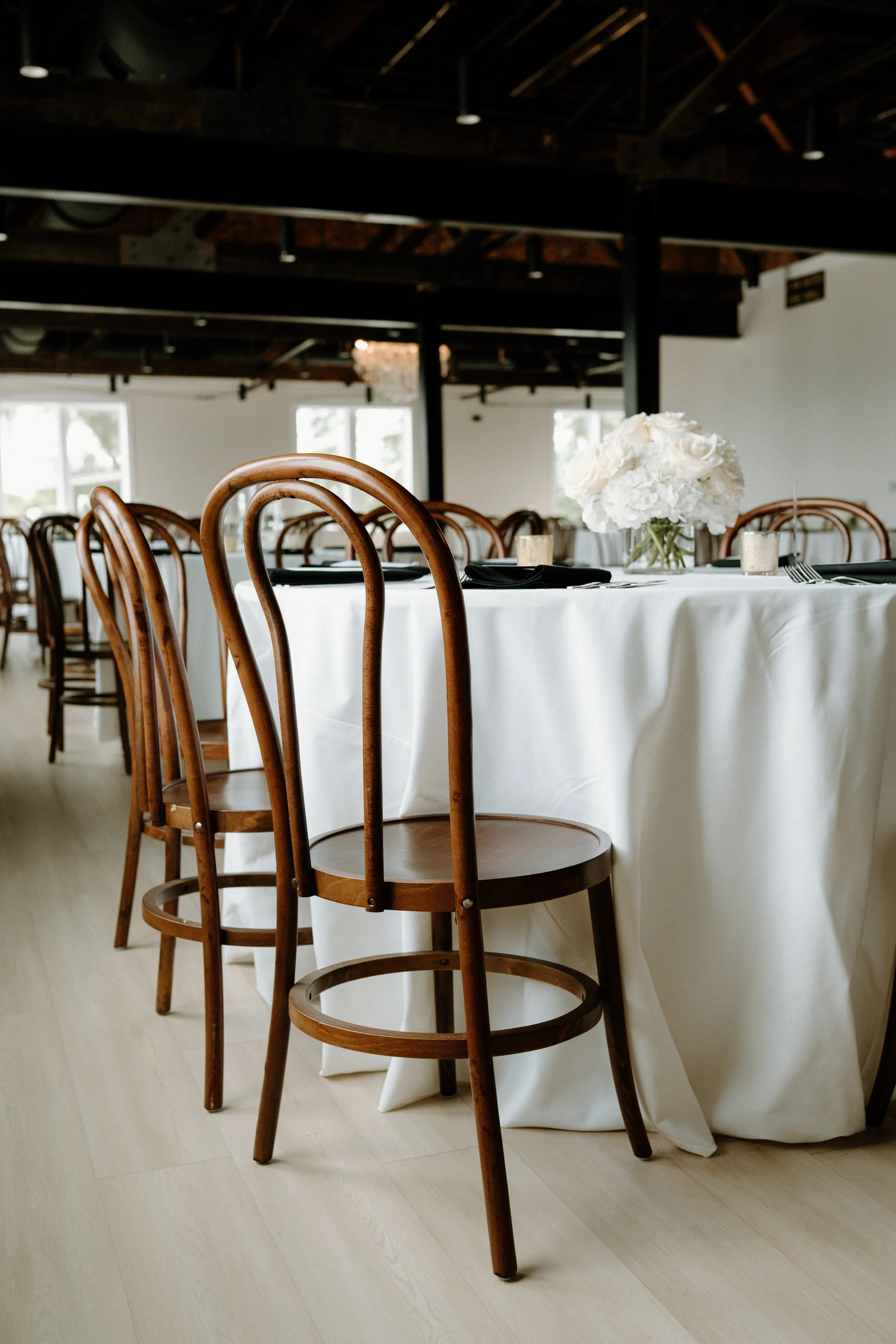 A round table with a white tablecloth, decorated with a flower arrangement, set for a formal event in a bright venue with wooden chairs and large windows.