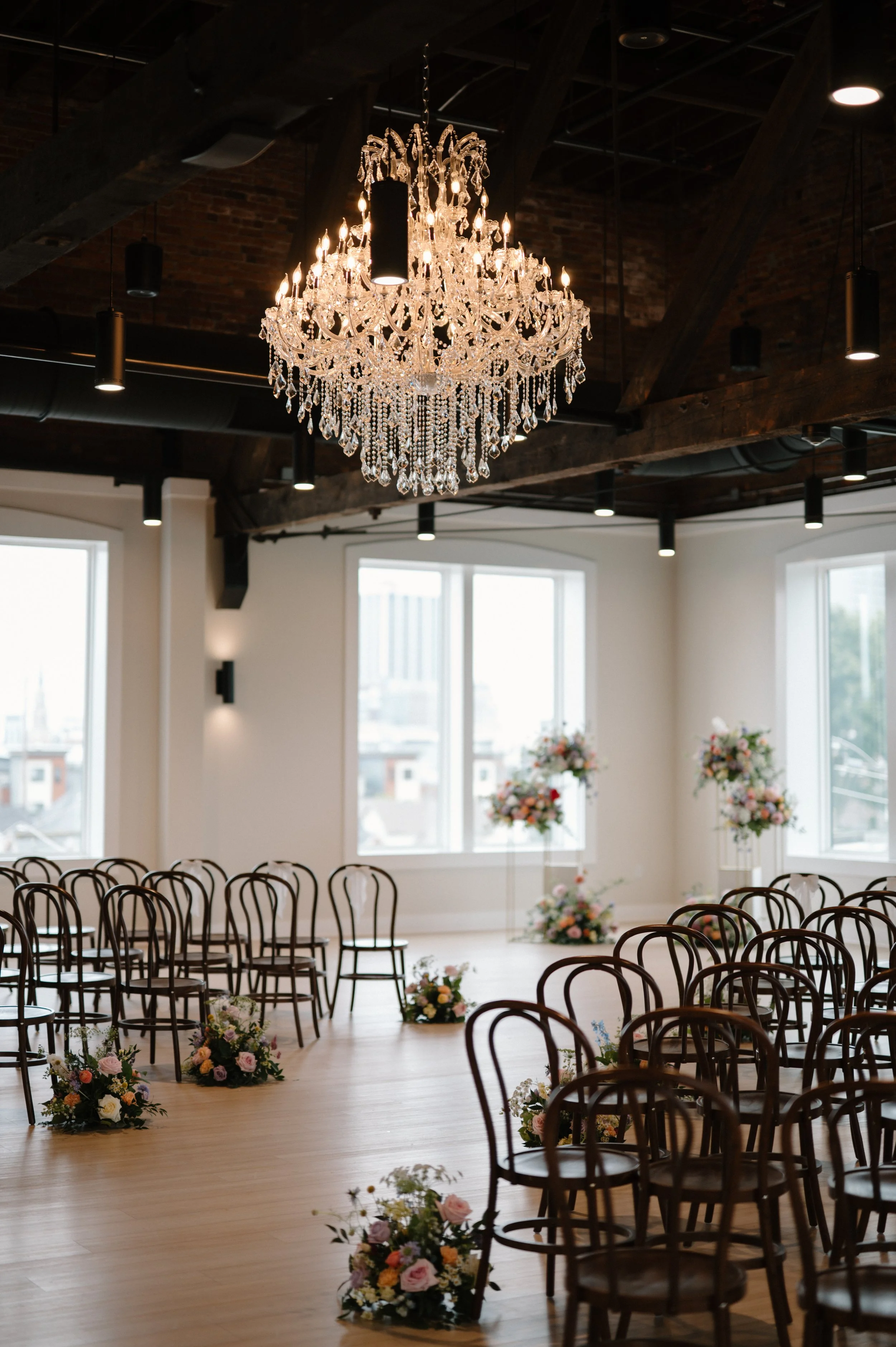 Elegant indoor wedding setup with a large crystal chandelier, wooden chairs arranged in rows, and floral decorations along the aisle in a bright room with large windows.