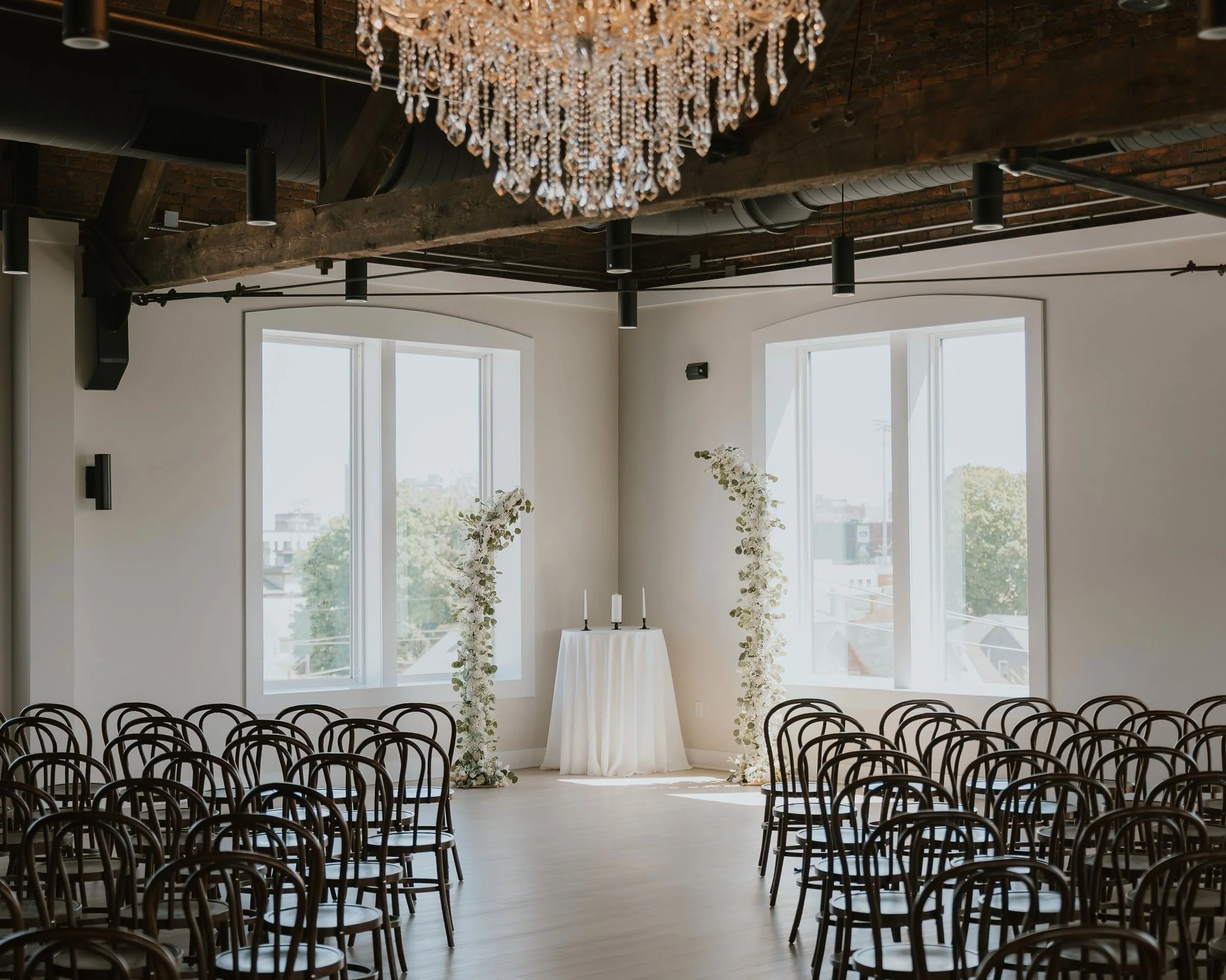 An indoor wedding ceremony setup with rows of wooden chairs facing a small altar with white flowers and candles, large windows letting in natural light, exposed wooden beams, and a sparkling chandelier overhead.