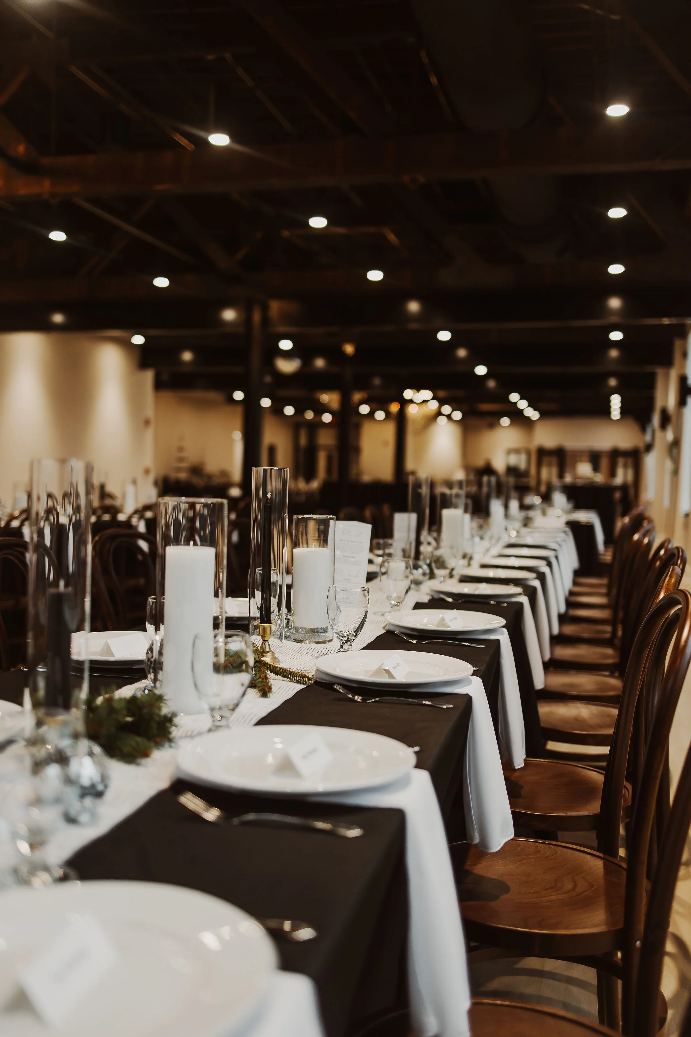 Elegant banquet table set with white plates, wine glasses, silverware, and tall glass candle holders in a decorated indoor venue with dark wood chairs and warm ceiling lighting.