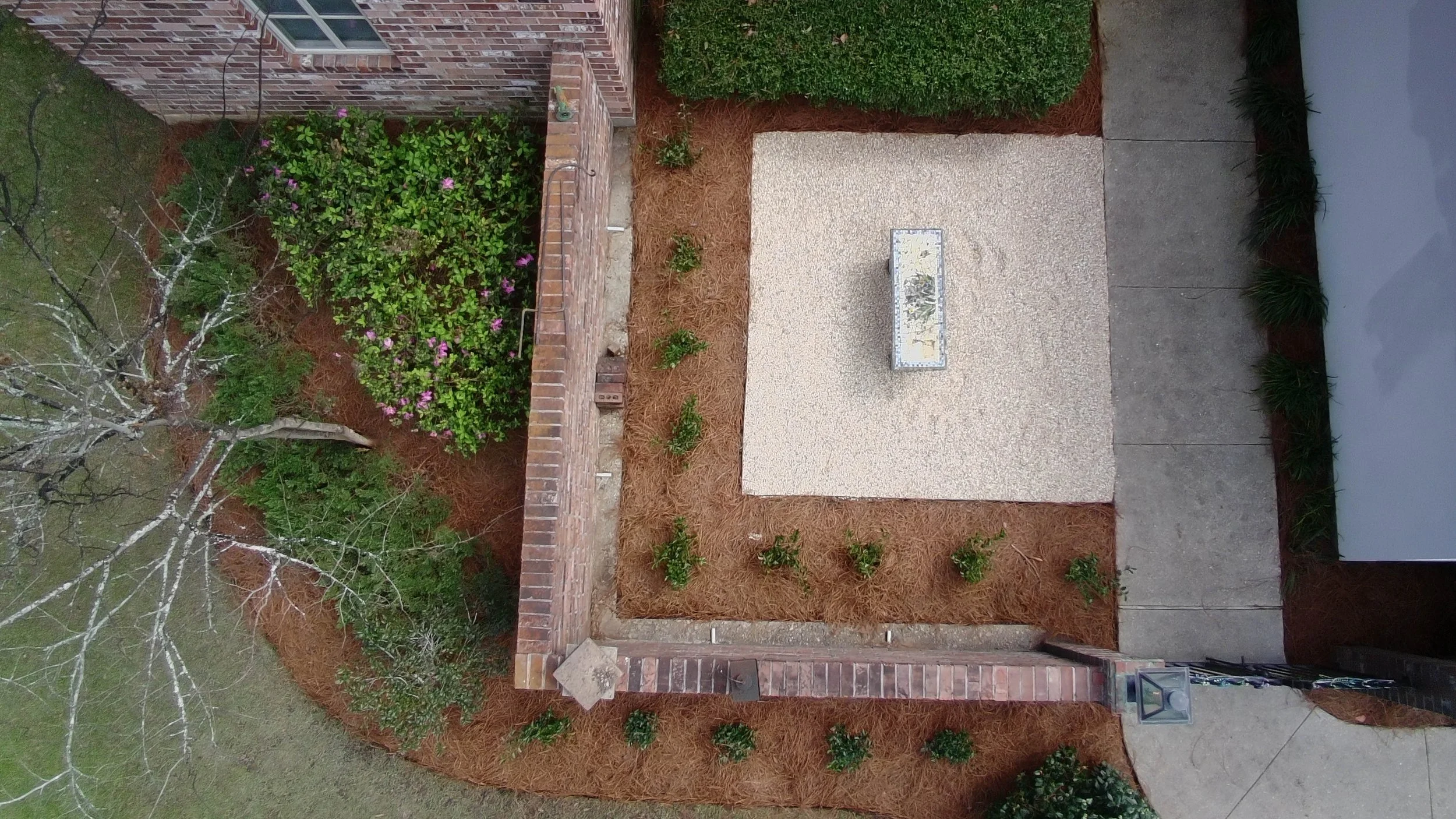 drone view of pea gravel rocky area. enclosed by a brick wall. garden bed wrapping around outside the brick wall with bushes