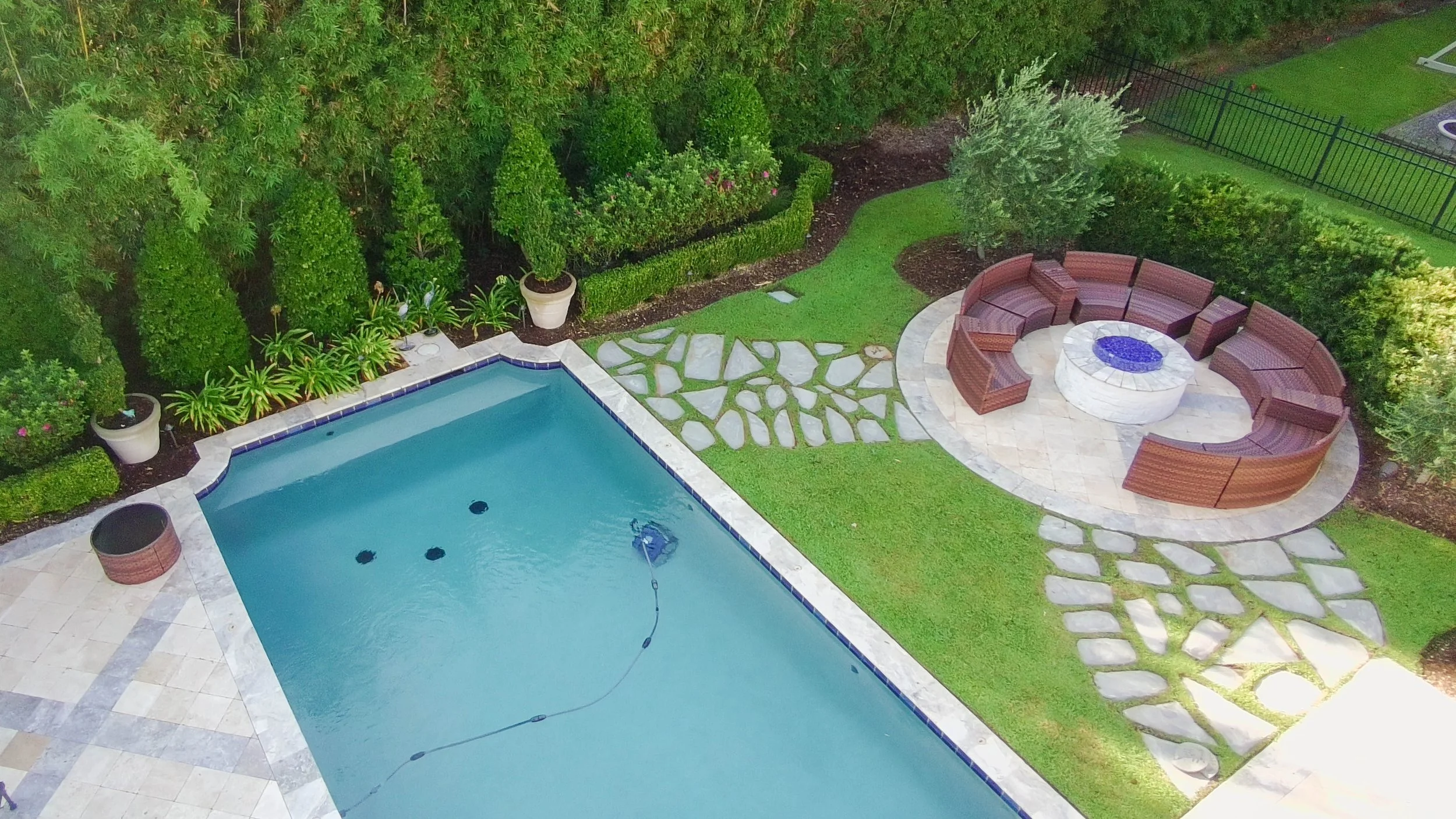 drone picture of a pool with landscaping lining the fence behind it & a paver walkway to a fire pit