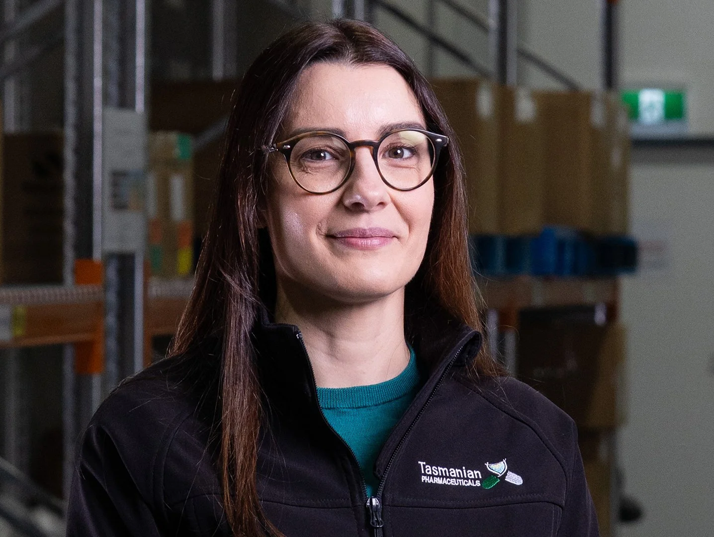 A woman with long brown hair and glasses, wearing a black Tasmanian Pharmaceuticals jacket, standing in a warehouse with shelves and boxes in the background.