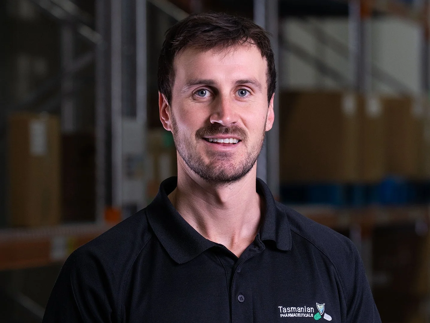 A man with brown hair and a beard smiling, wearing a black collared shirt with a logo that reads 'Tasmanian Pharmaceutical' in an industrial warehouse setting with shelves and boxes in the background.