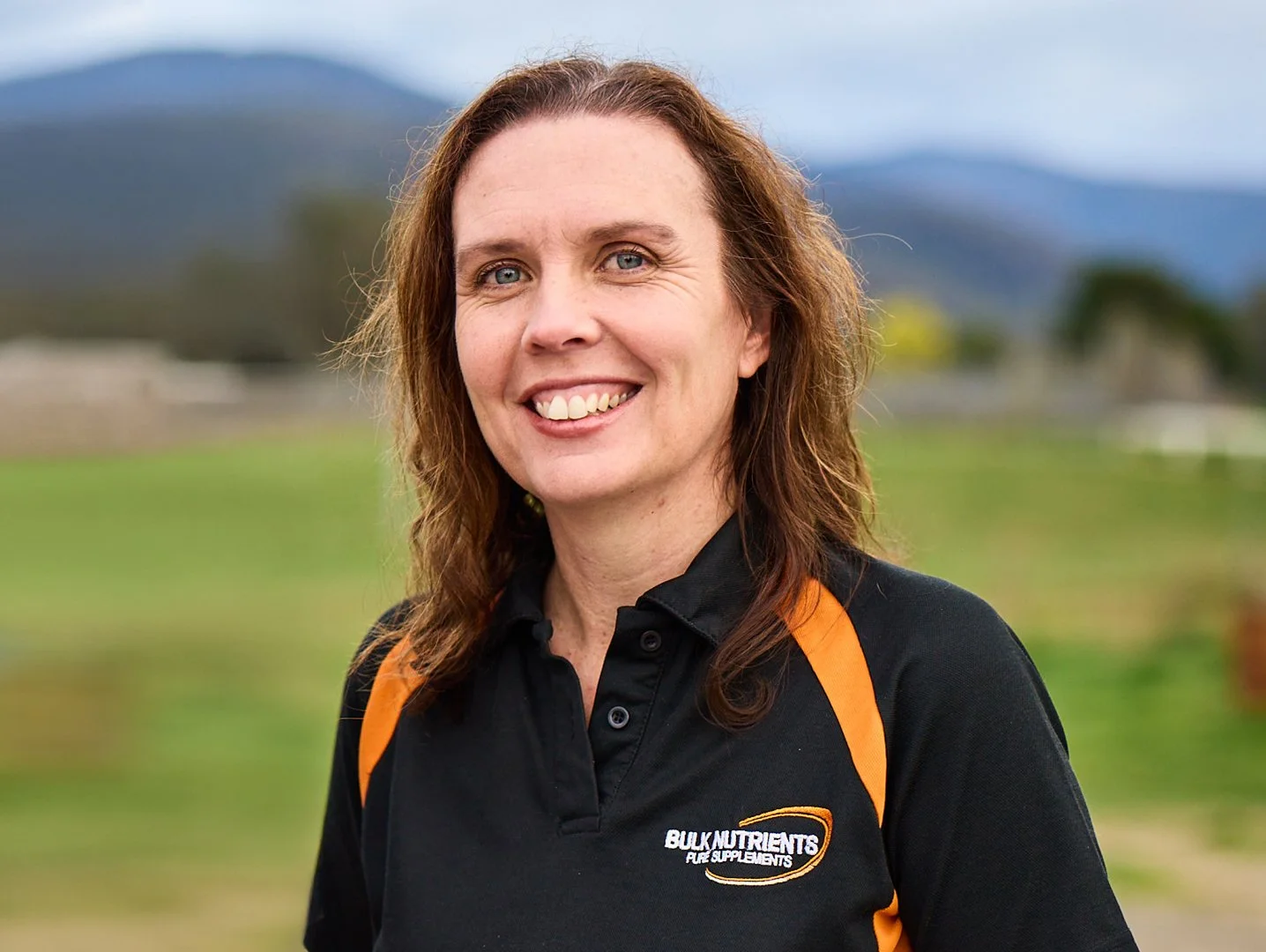A woman with long, curly red hair smiling outdoors, wearing a black polo shirt with orange accents and a logo for Bulk Nutrients.