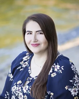 A woman with long brown hair, winking and smiling, wearing a navy blue floral blouse, outdoors in front of a blurred green and yellow background.