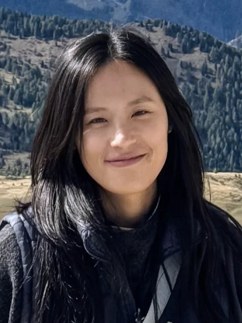 Young woman with long dark hair smiling outdoors with mountains and trees in the background.
