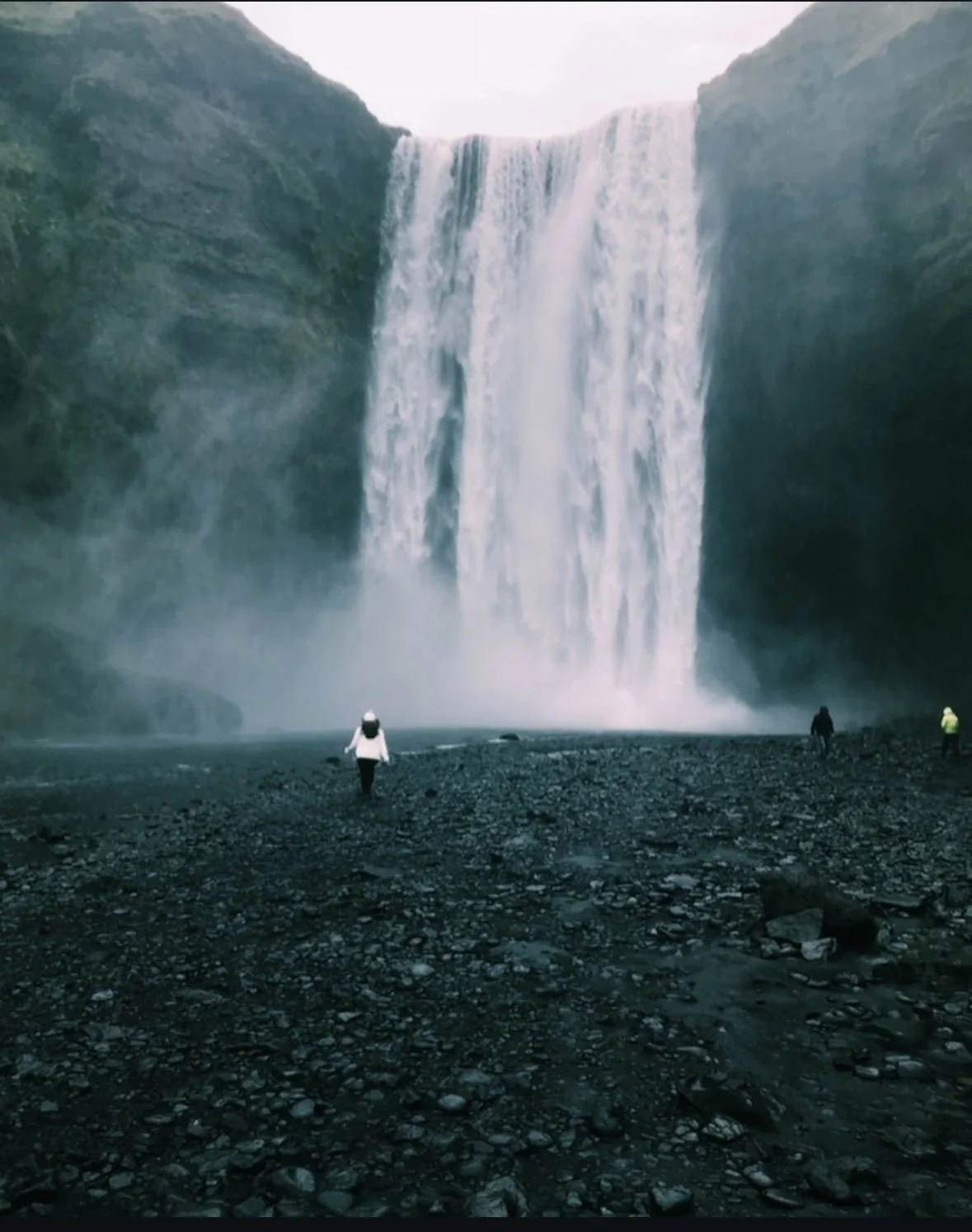 People standing on rocky ground in front of a large waterfall surrounded by green cliffs.