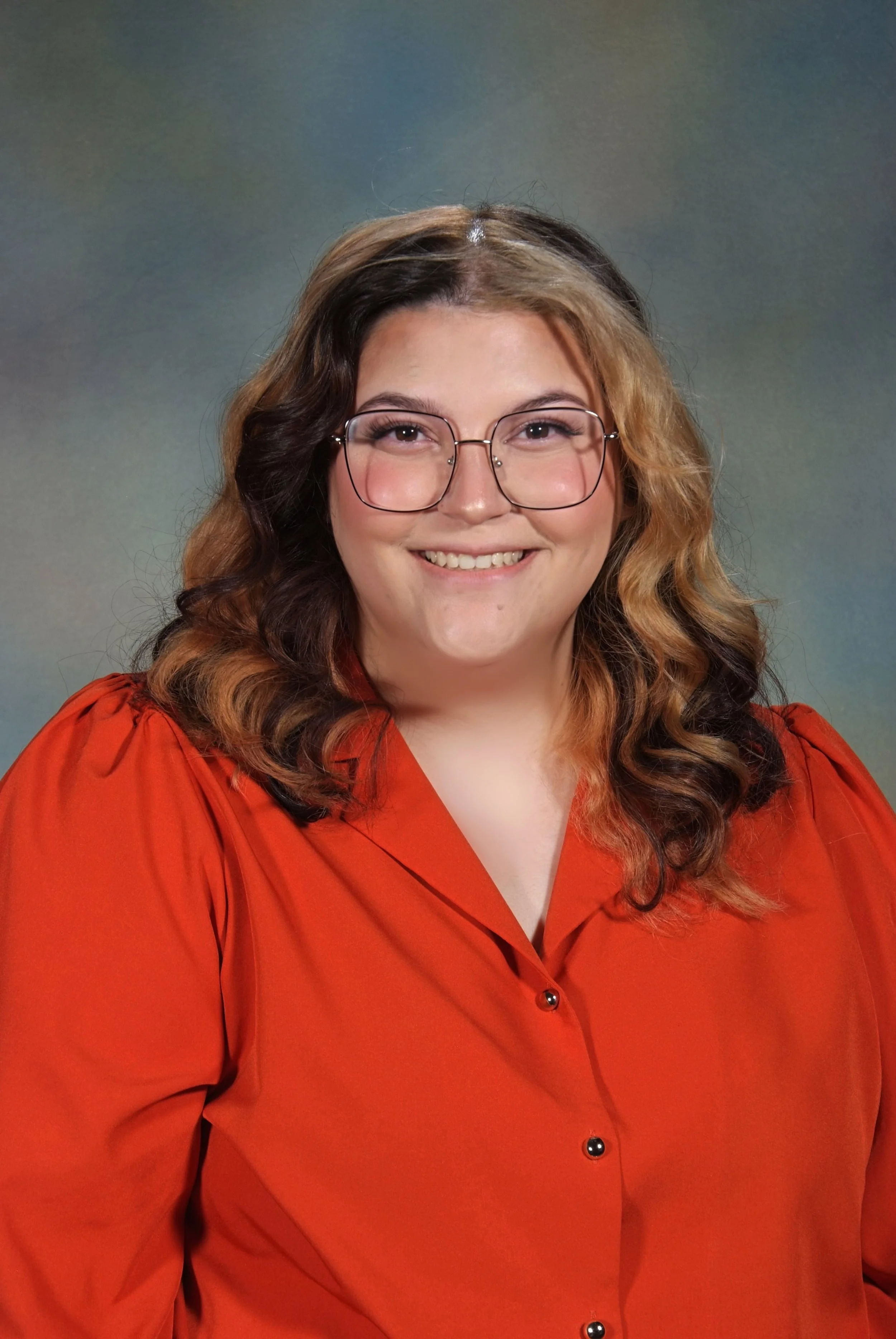 A young woman with glasses and wavy hair is smiling and wearing an orange shirt, posing against a multicolored background.