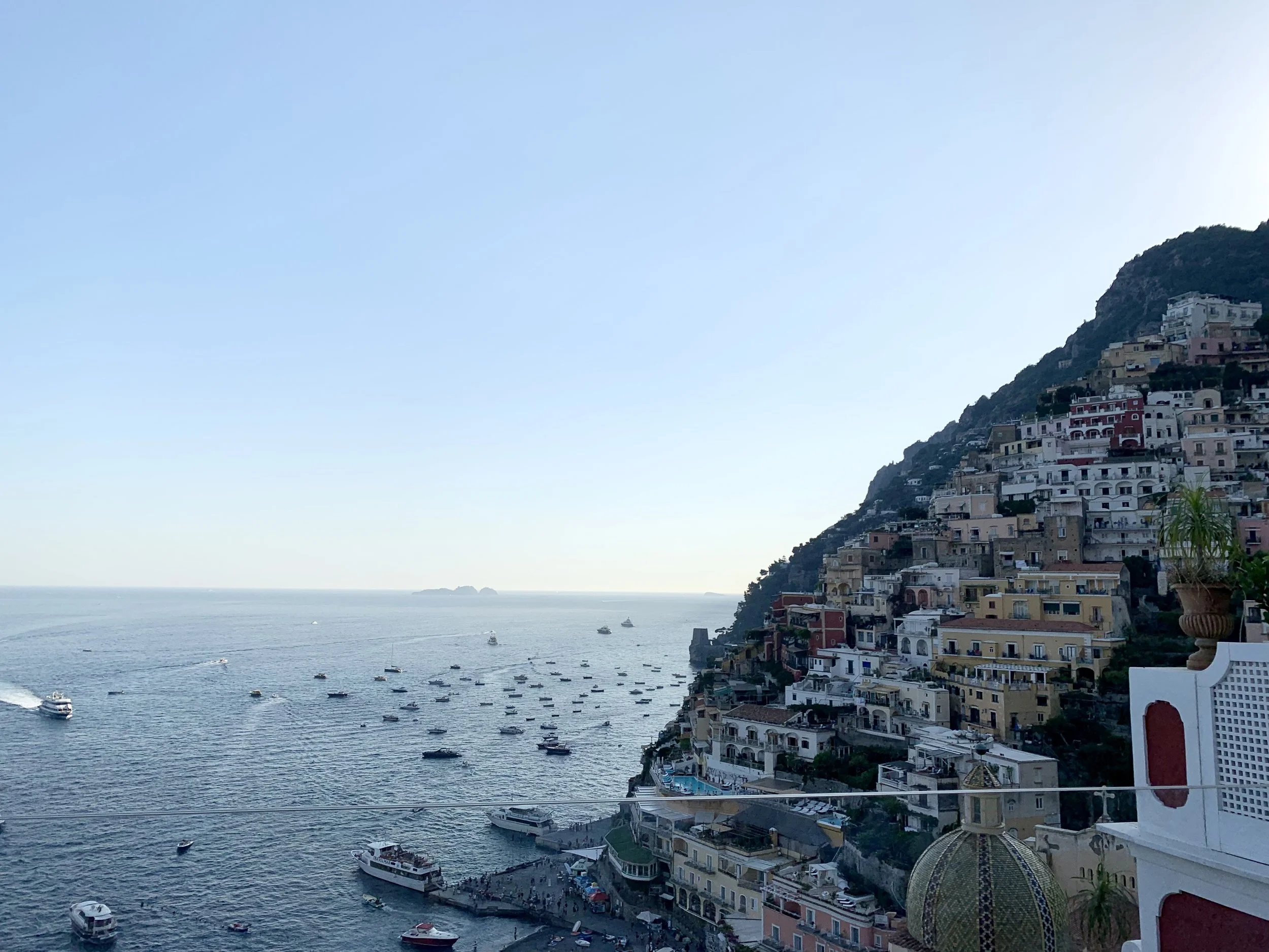 Scenic view of a coastal hillside town with numerous colorful buildings, overlooking the ocean with boats and a small island in the distance.