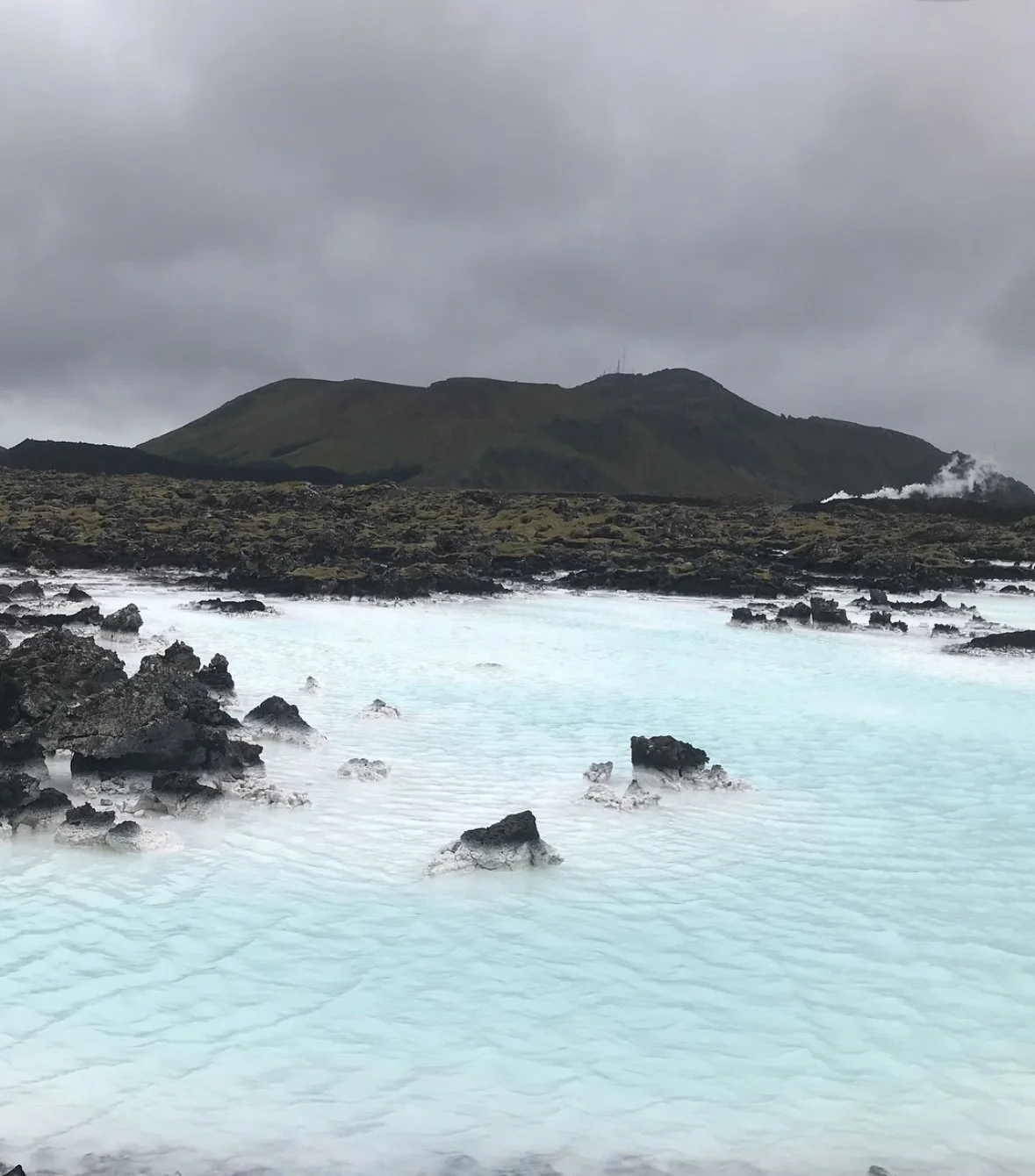 A geothermal hot spring with milky blue water surrounded by black volcanic rocks, with a green mountain in the background under a cloudy sky.