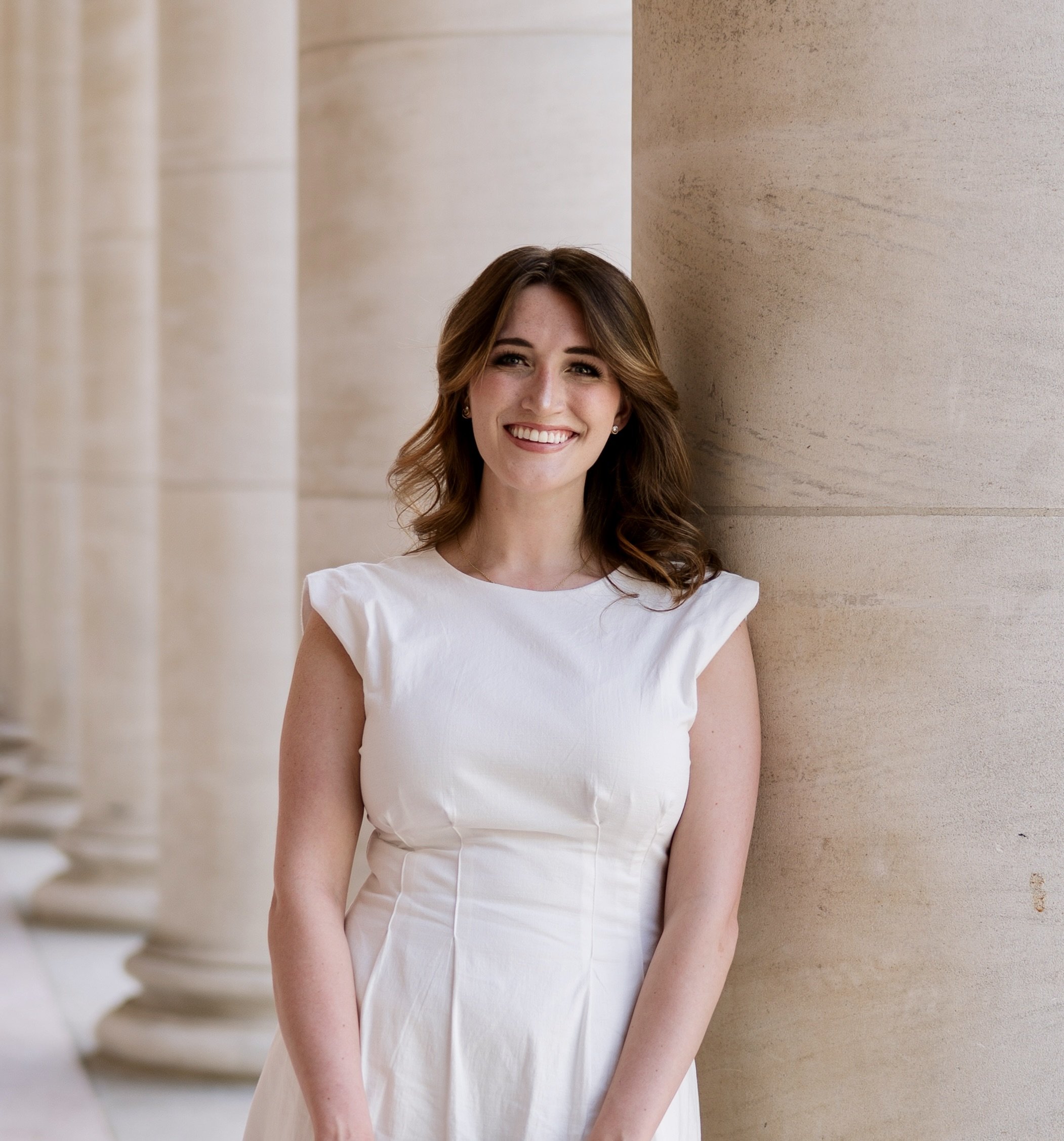 A young woman with wavy brown hair and fair skin smiling, wearing a white dress, standing against a beige stone or concrete wall with architectural columns on the left side.
