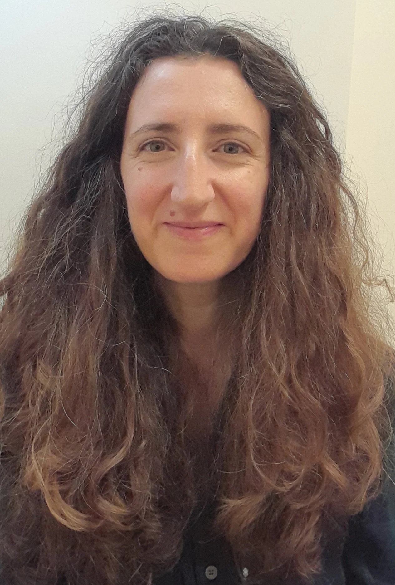 A woman with long, curly brown hair smiling at the camera, wearing a black top, against a plain beige background.