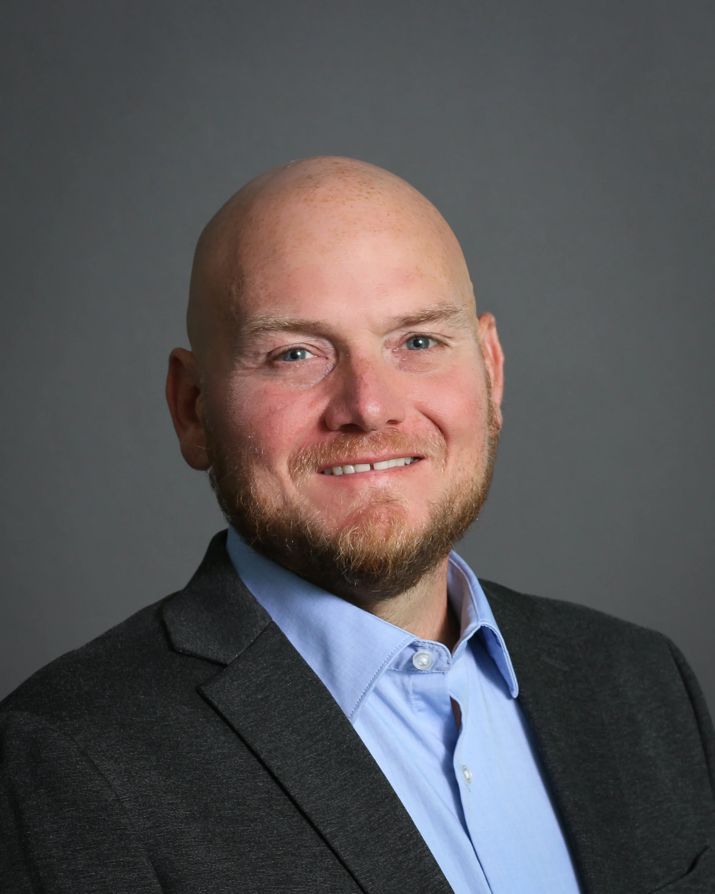 Close-up of a smiling man with a shaved head, blue eyes, and a beard, wearing a dark gray suit jacket and a light blue collared shirt, against a plain gray background.