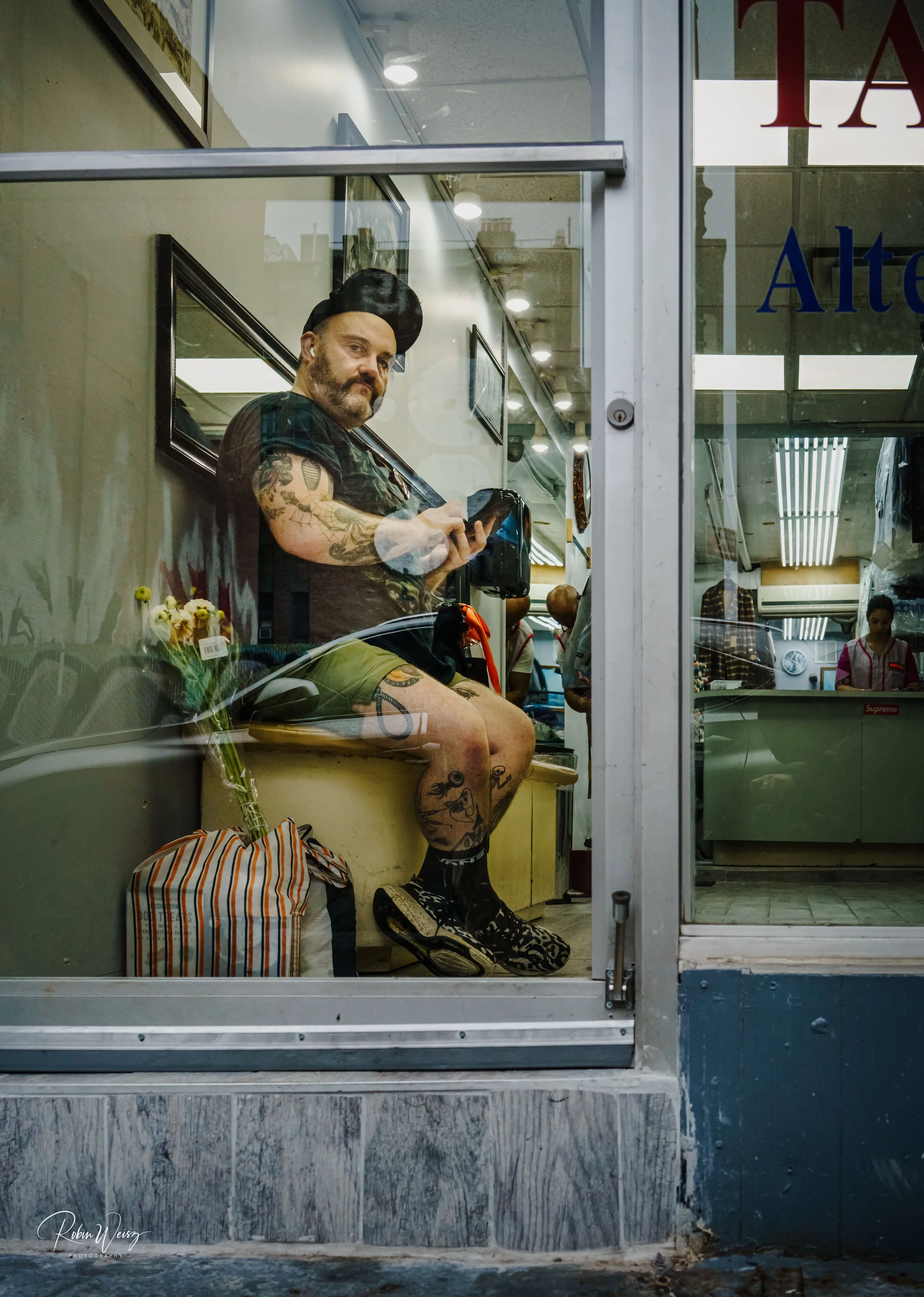 Tattooed barber wearing a black cap cuts hair inside a barbershop, viewed through the storefront window.