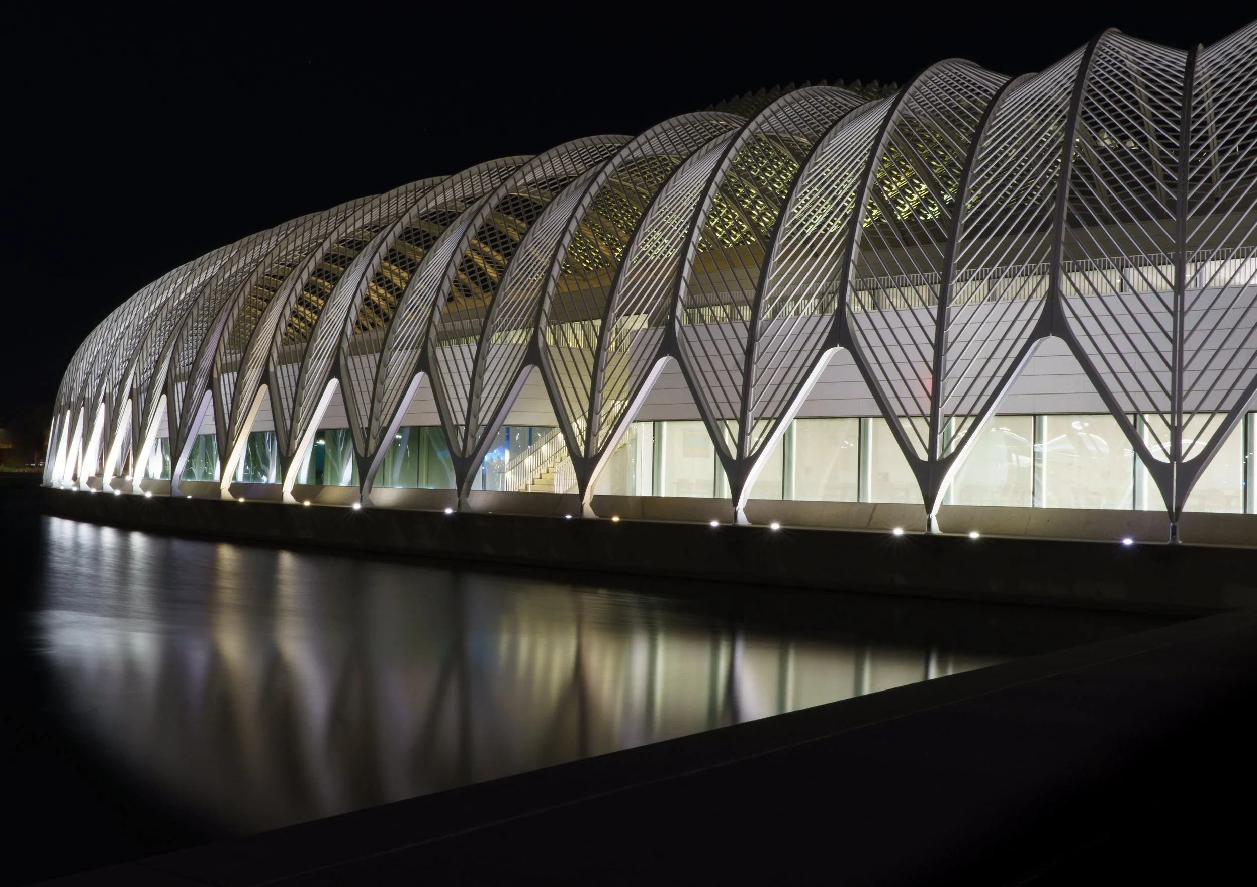 Illuminated modern architectural building with repeating curved steel arches reflected in a waterfront at night
