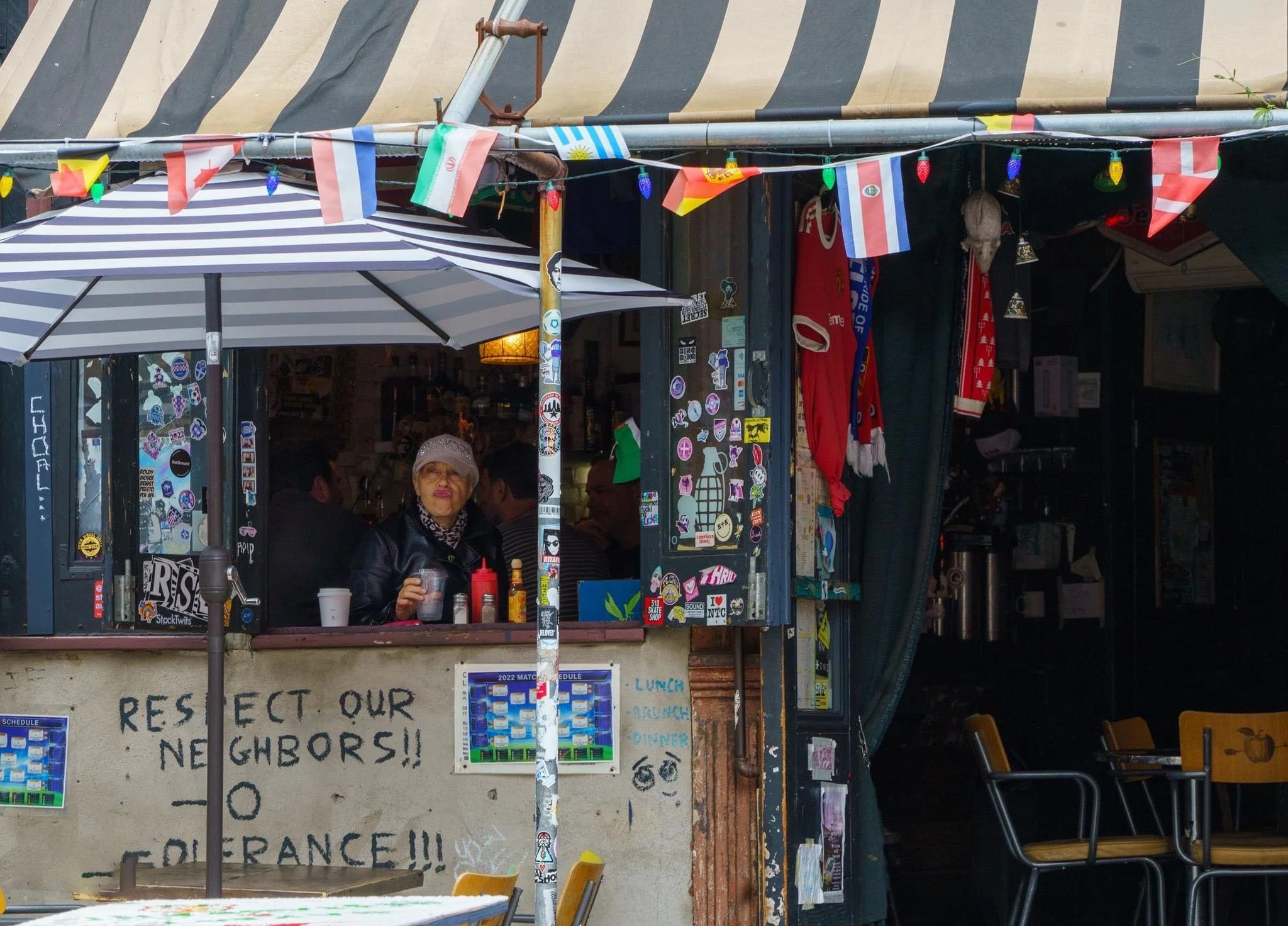 Street cafe storefront decorated with international flags, string lights, umbrellas, sports scarves, and sticker-covered walls