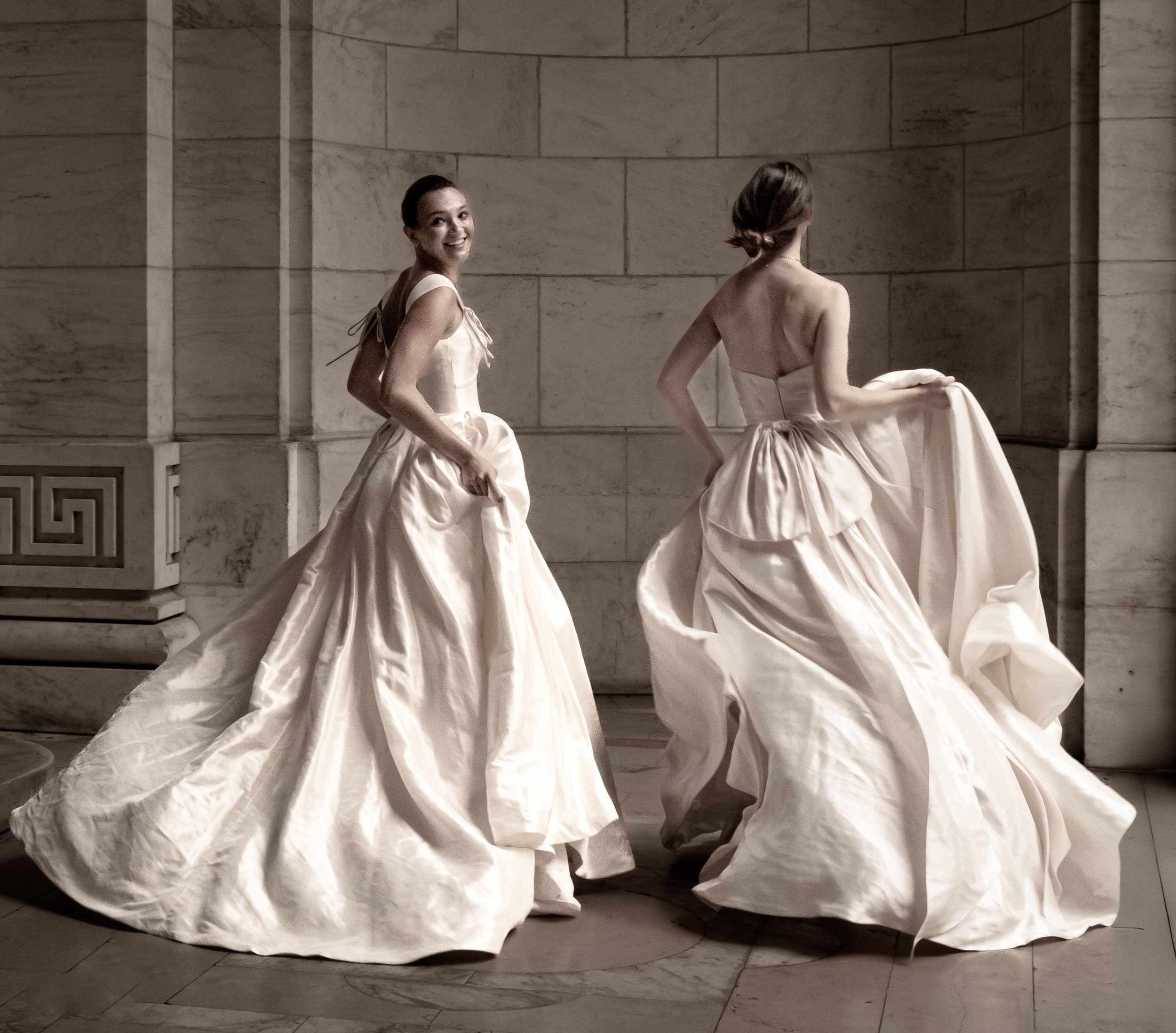 Two women in flowing white gowns pose against a stone wall, one smiling at the camera and the other facing away.