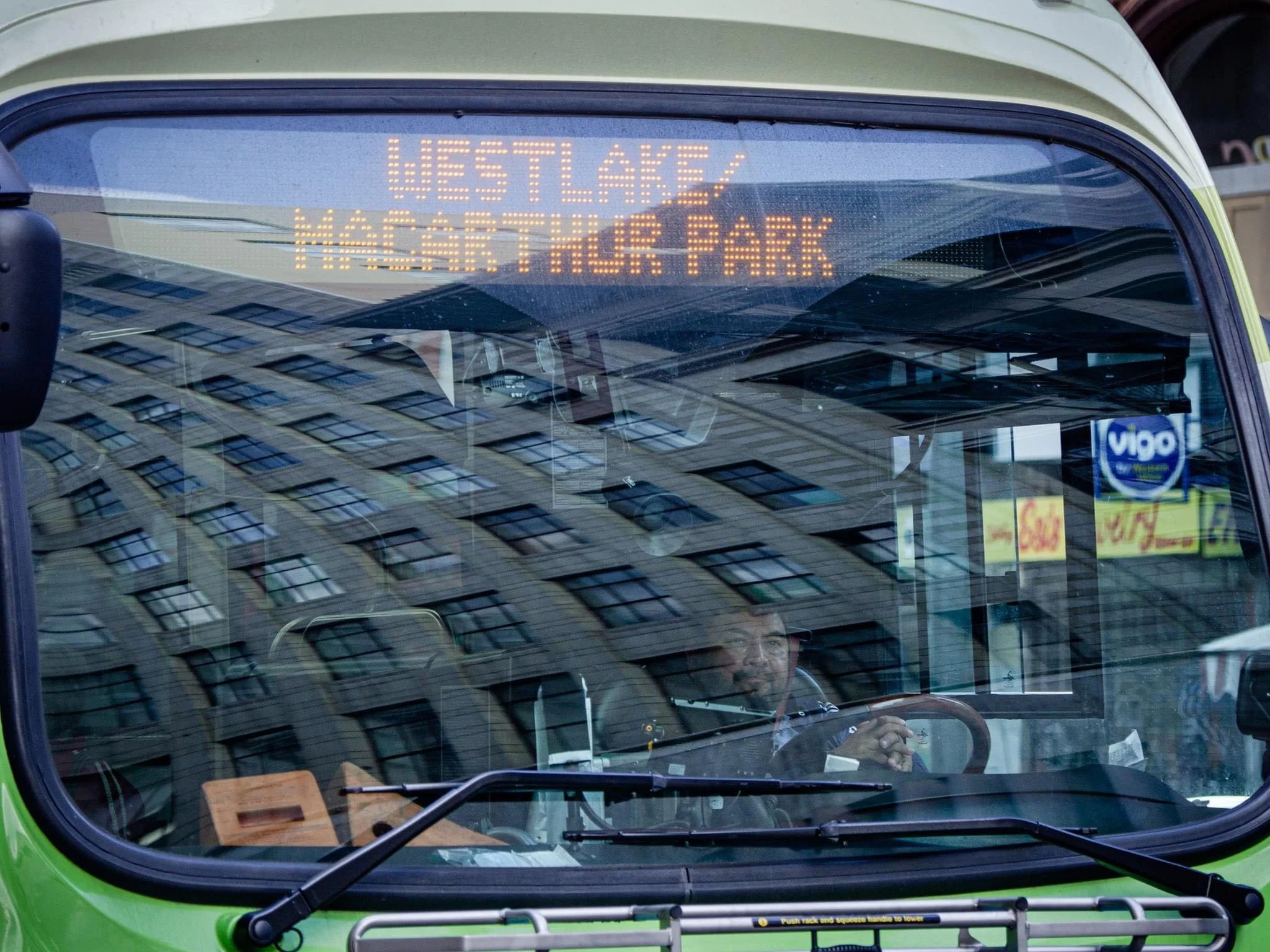 Front of a green city bus displaying “DESTLAKE” and “NORTH HARBOUR” on its LED destination sign, with the driver visible through the windshield.