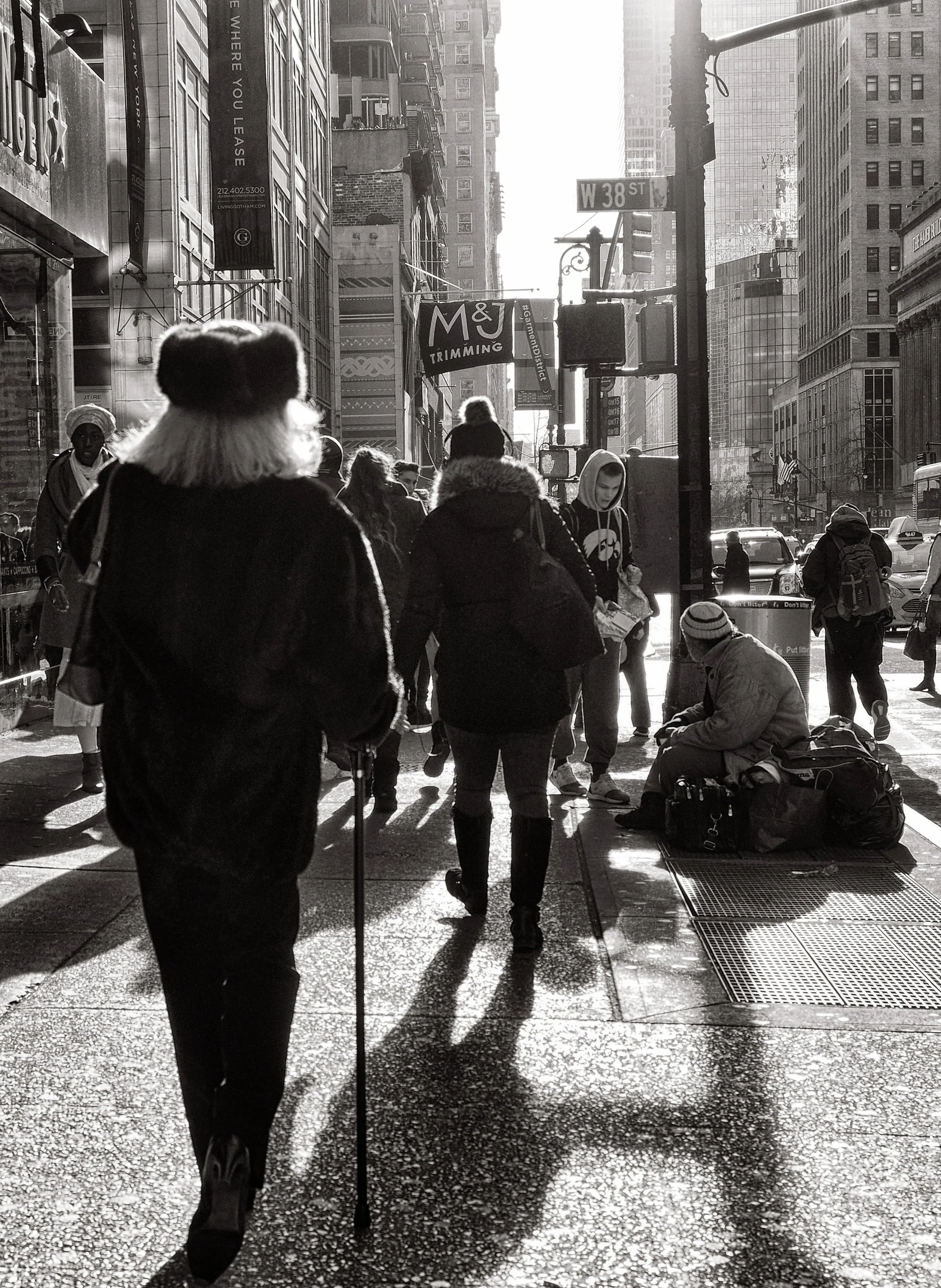 Black-and-white street scene at West 38th Street in New York City, with pedestrians, taxis, skyscrapers, and a homeless person near a subway grate.