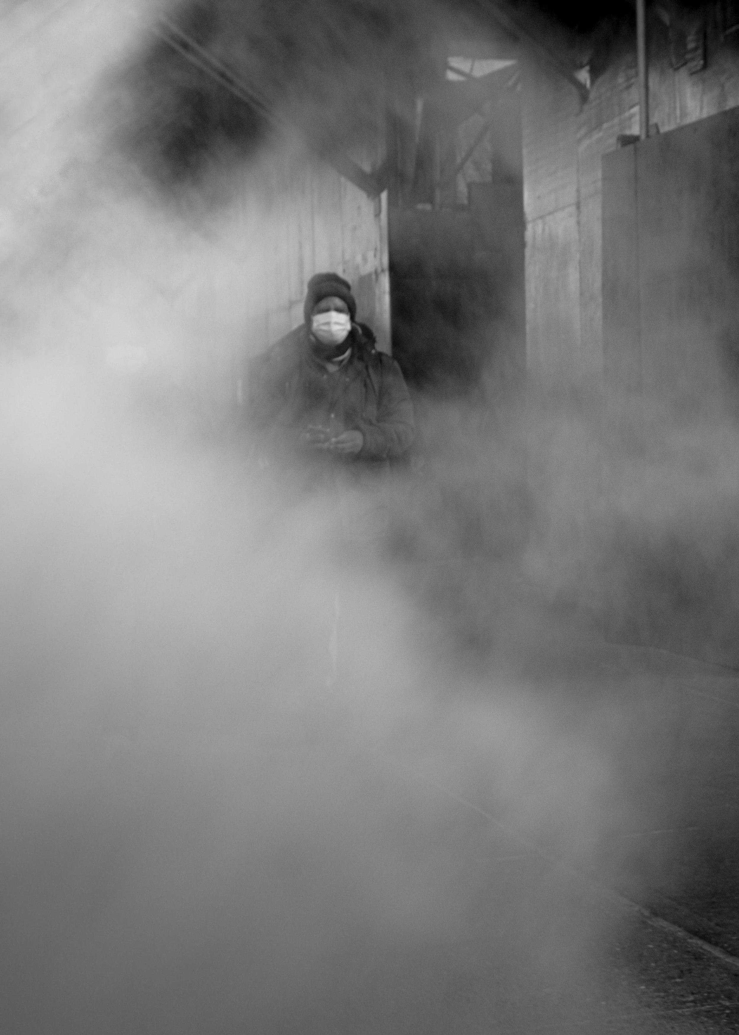 Person wearing a knit hat, winter coat, and face mask walking through a foggy industrial alley.