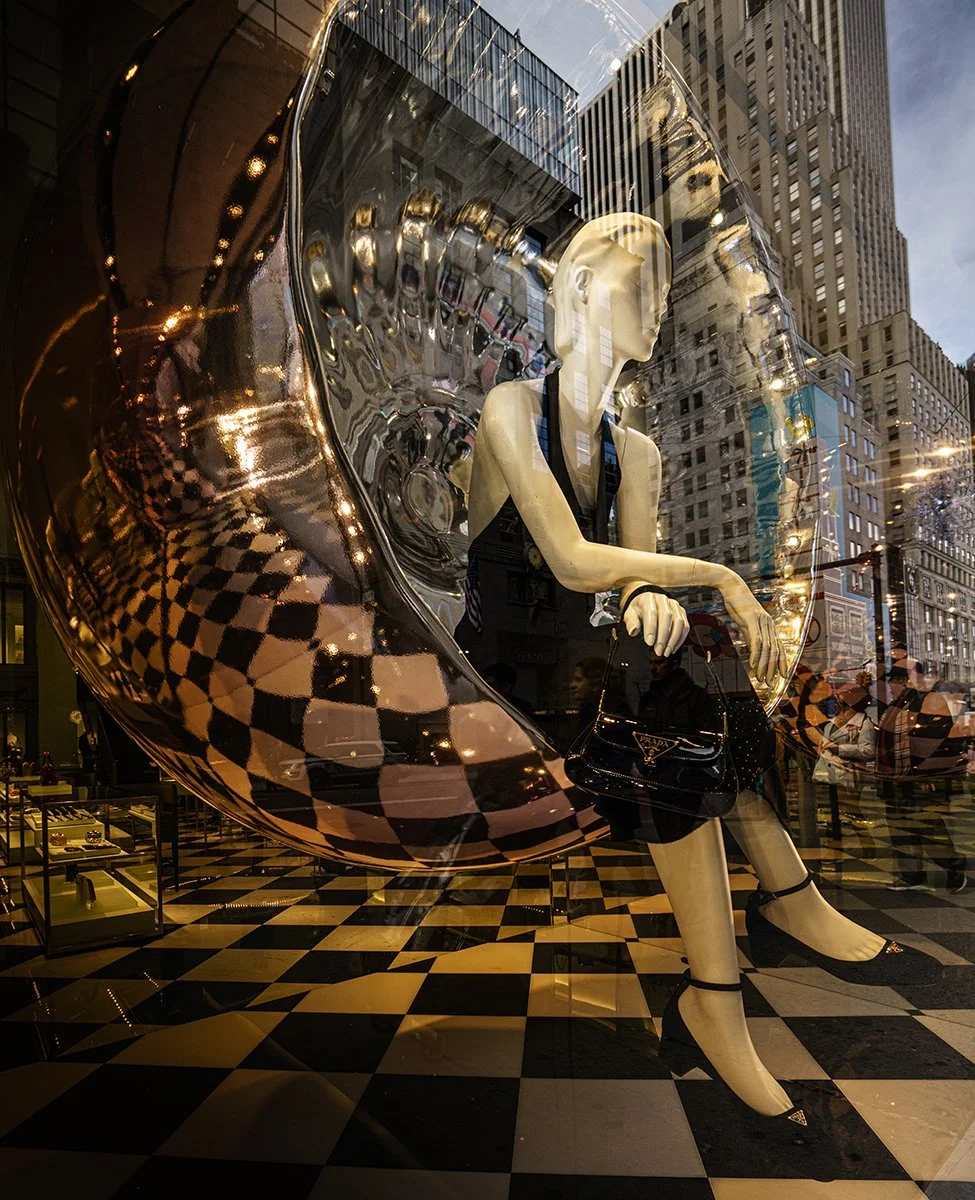 Prada store window display with a mannequin beside a giant metallic high-heel sculpture and black-and-white checkerboard floor in a city shopping district.