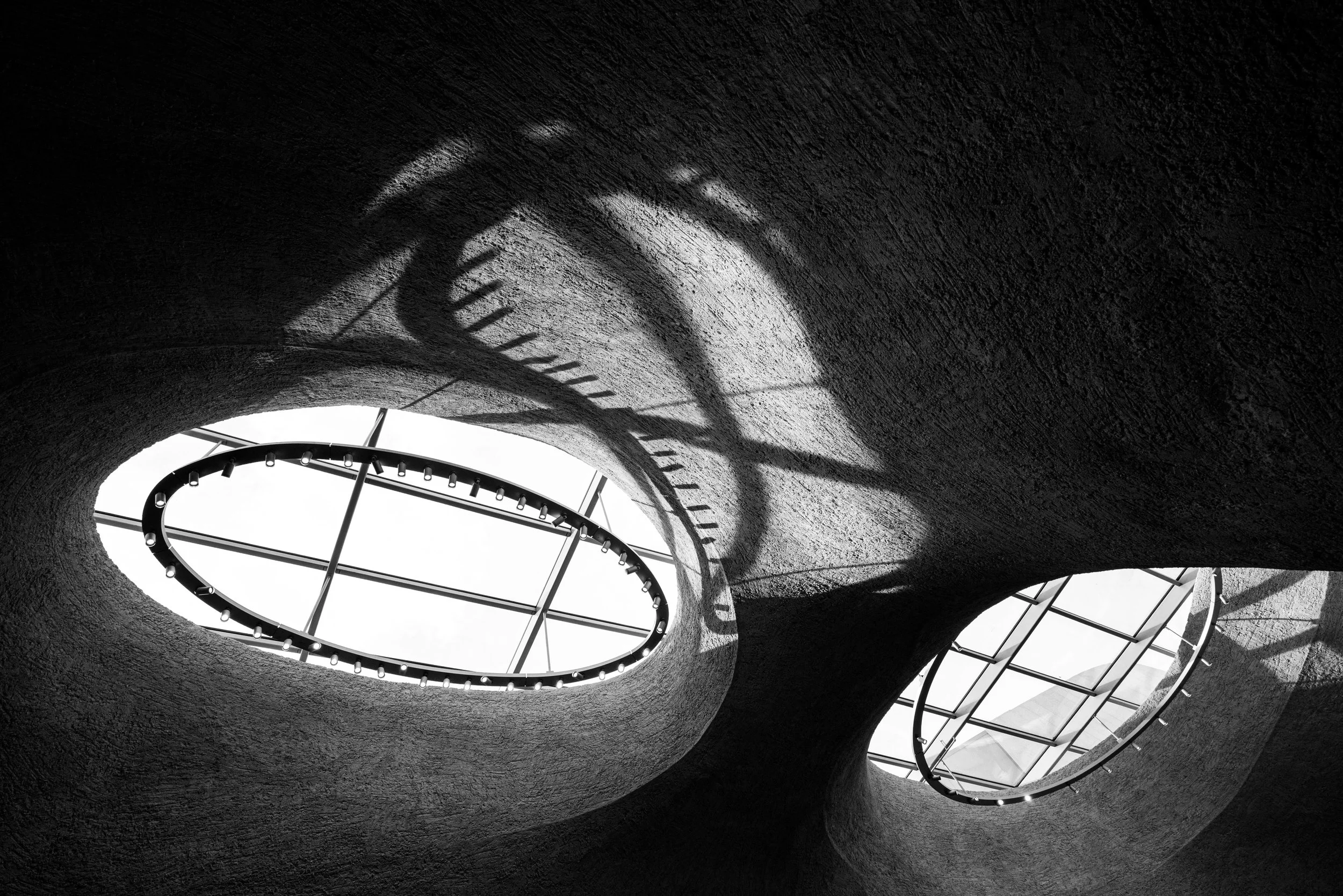Black-and-white upward view of a sculptural concrete structure with oval skylights, metal frames, and ring lights