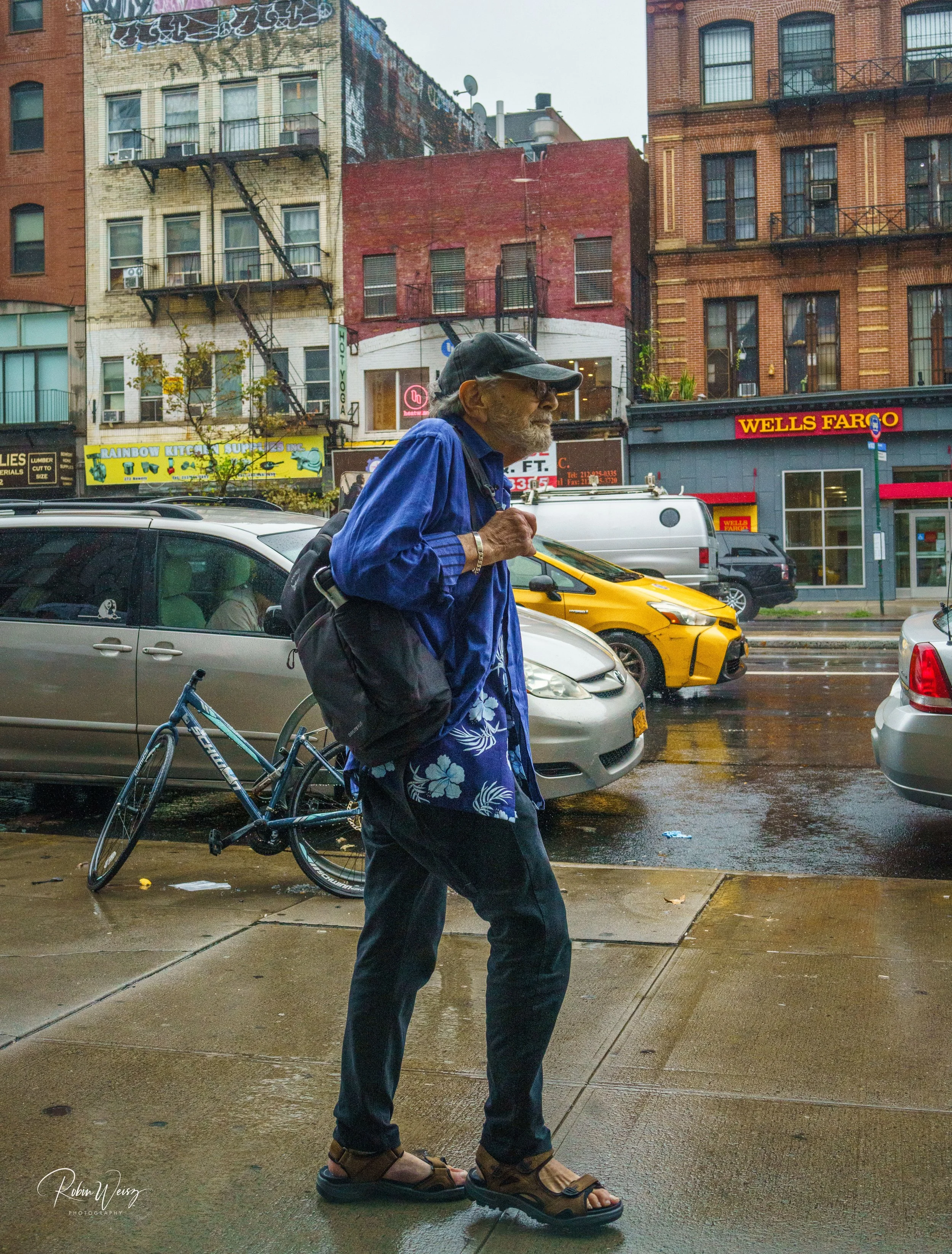 Older man in a blue shirt and black cap walking with a bicycle on a rainy city sidewalk, with taxis, storefronts, brick buildings, and a Wells Fargo bank behind him.