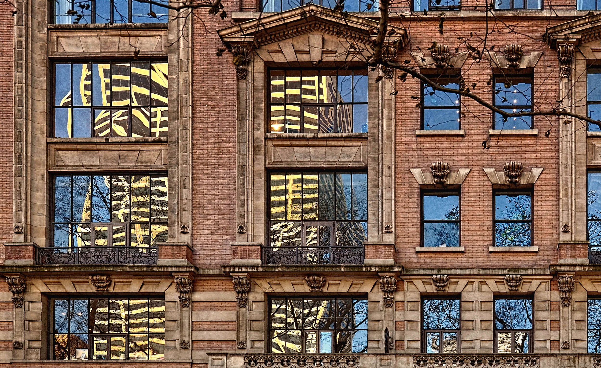 Close-up of an ornate historic brick building facade with large reflective windows, stone detailing, and bare tree branches.