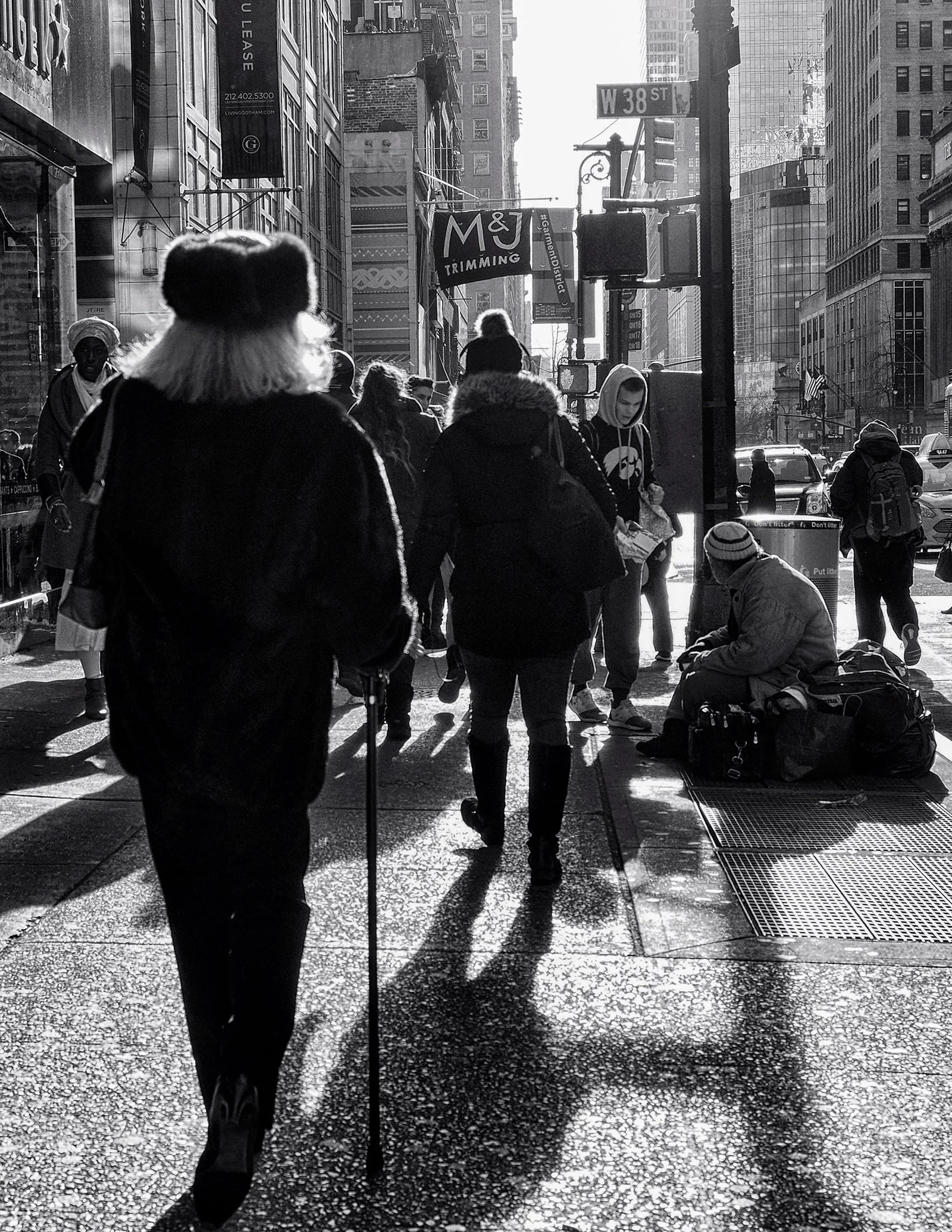 Black-and-white street scene at West 38th Street in Manhattan, with pedestrians, taxis, skyscrapers, and a seated person with belongings.