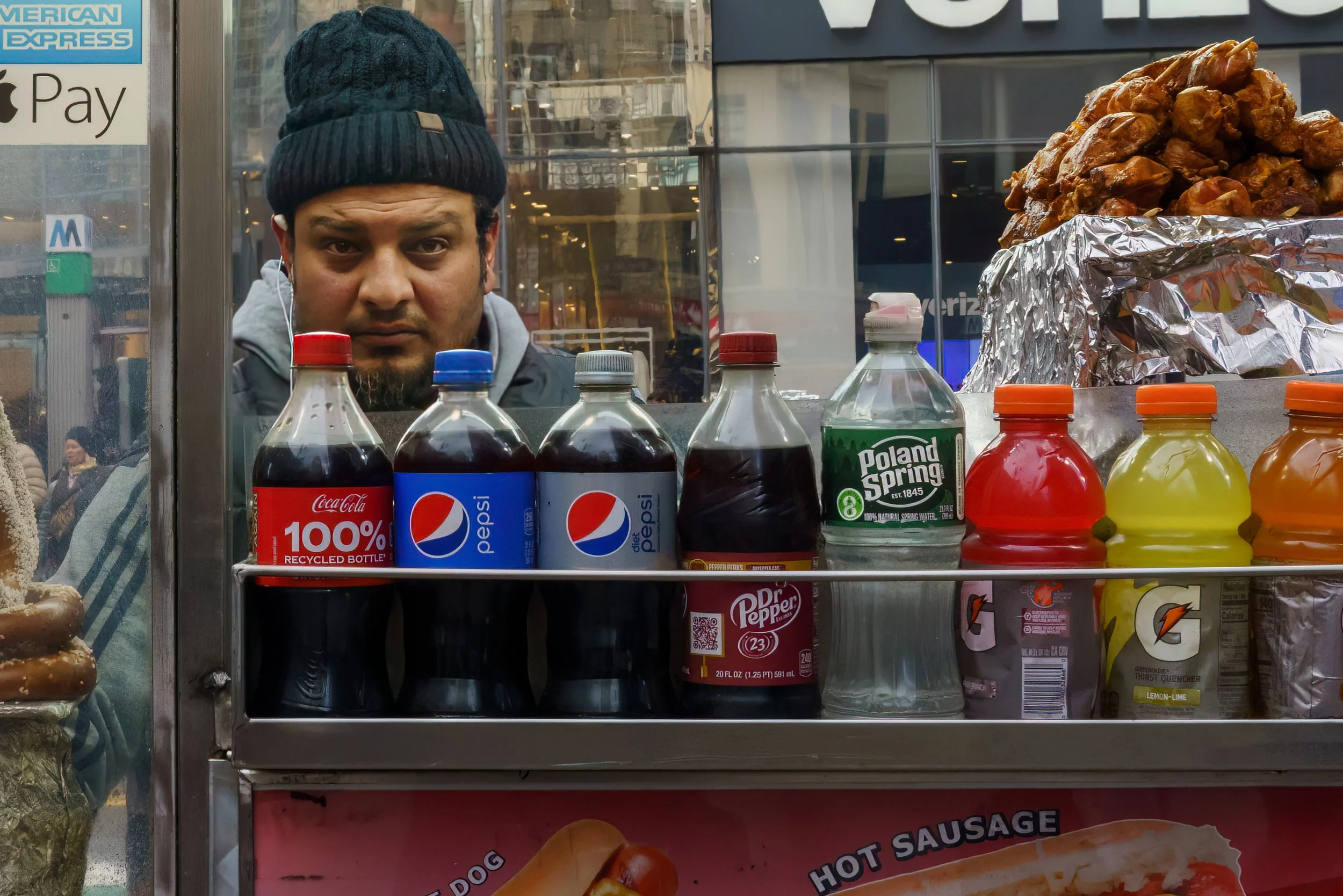 Street food cart displaying assorted sodas and fried snacks, with a man reflected in the window behind it