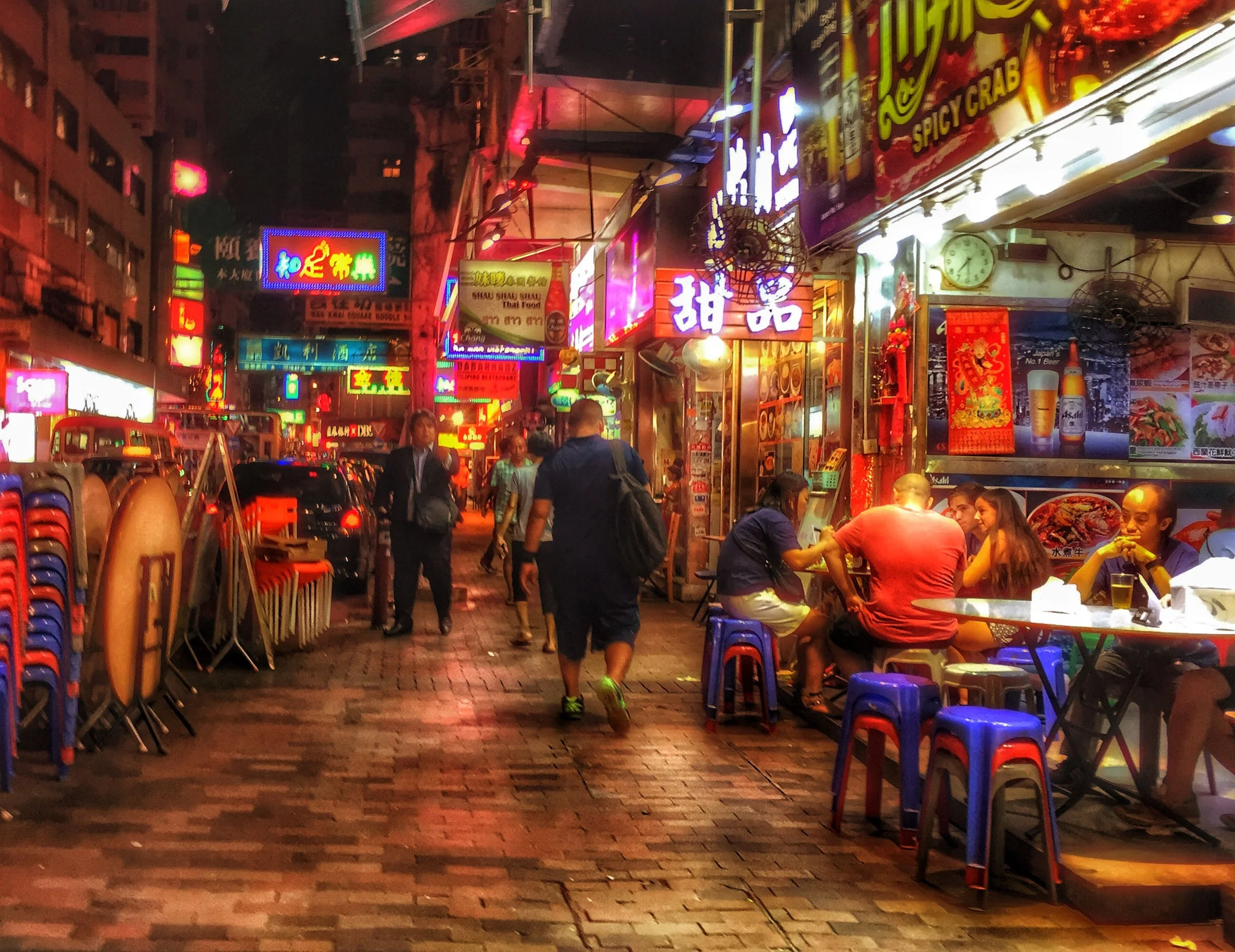Busy nighttime street in Hong Kong lined with brightly lit Chinese restaurant signs, outdoor dining tables, pedestrians, and colorful plastic stools.