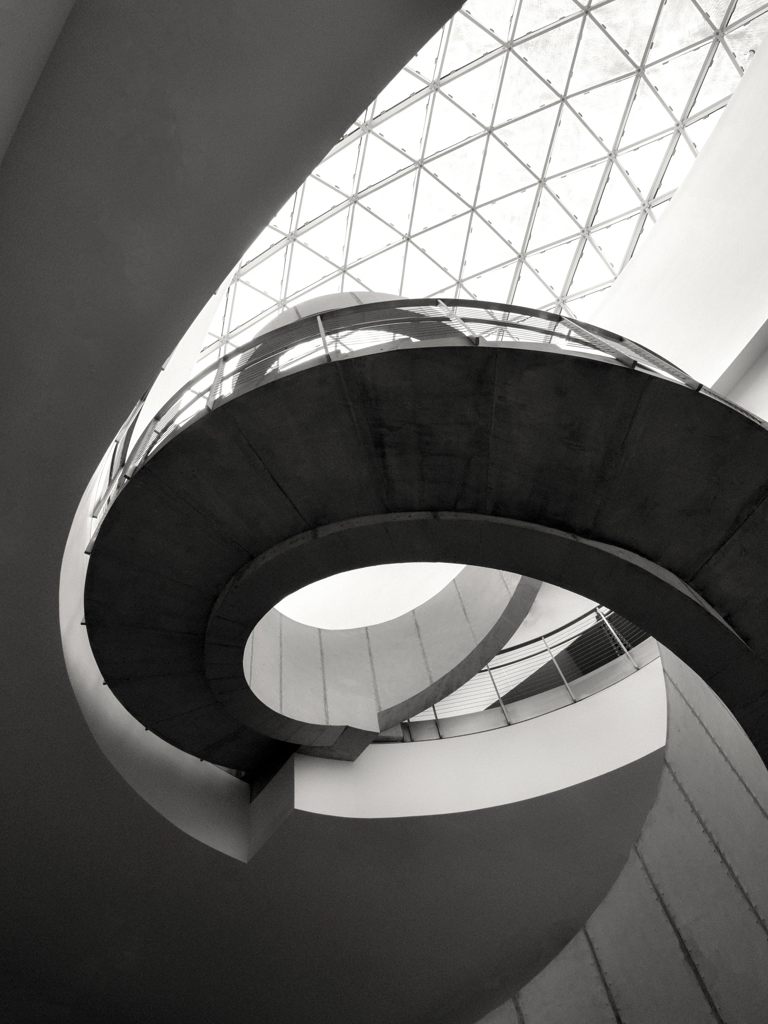 Black-and-white upward view of a modern atrium with a geometric glass dome, curved architectural balcony, and sweeping structural arches.