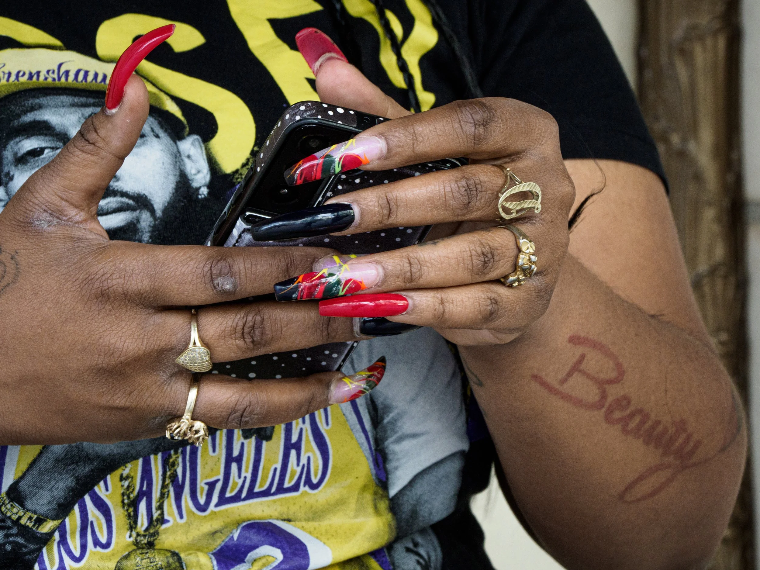 Close-up of a person with long colorful acrylic nails holding a black smartphone and wearing gold rings.