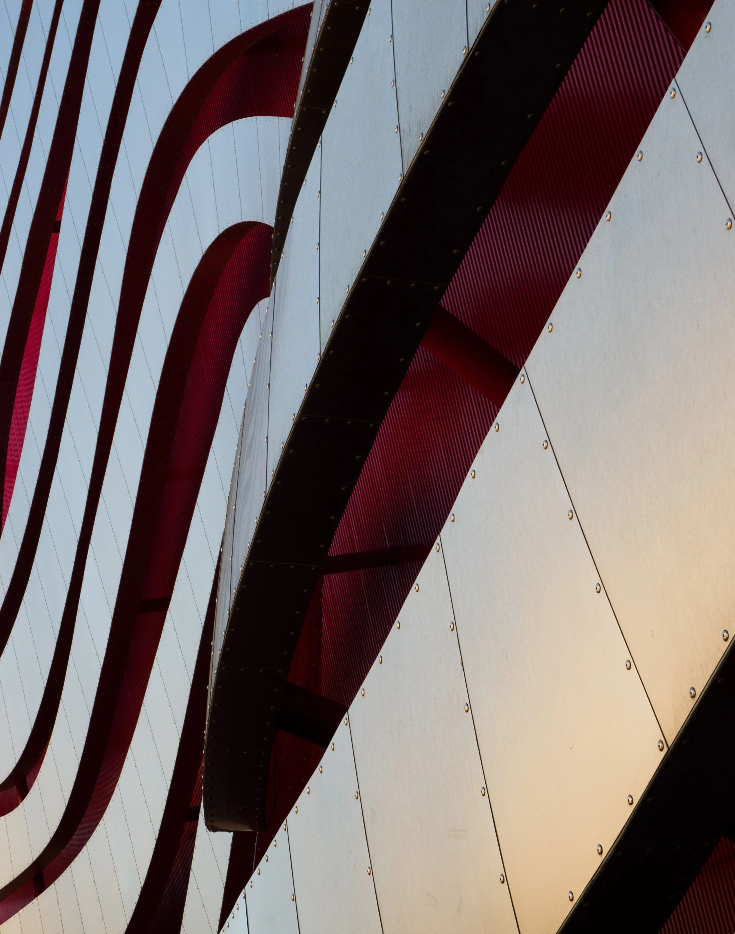 Abstract close-up of curved red and black metal beams against riveted silver panels and blue sky
